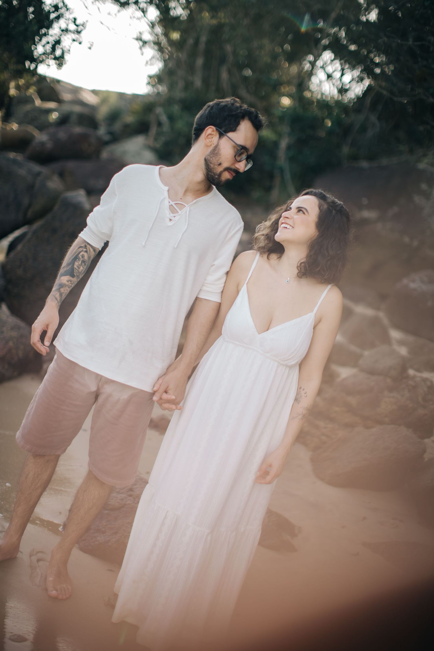 Romantic couple in coordinated white outfits sharing a joyful moment on a rocky beach.
