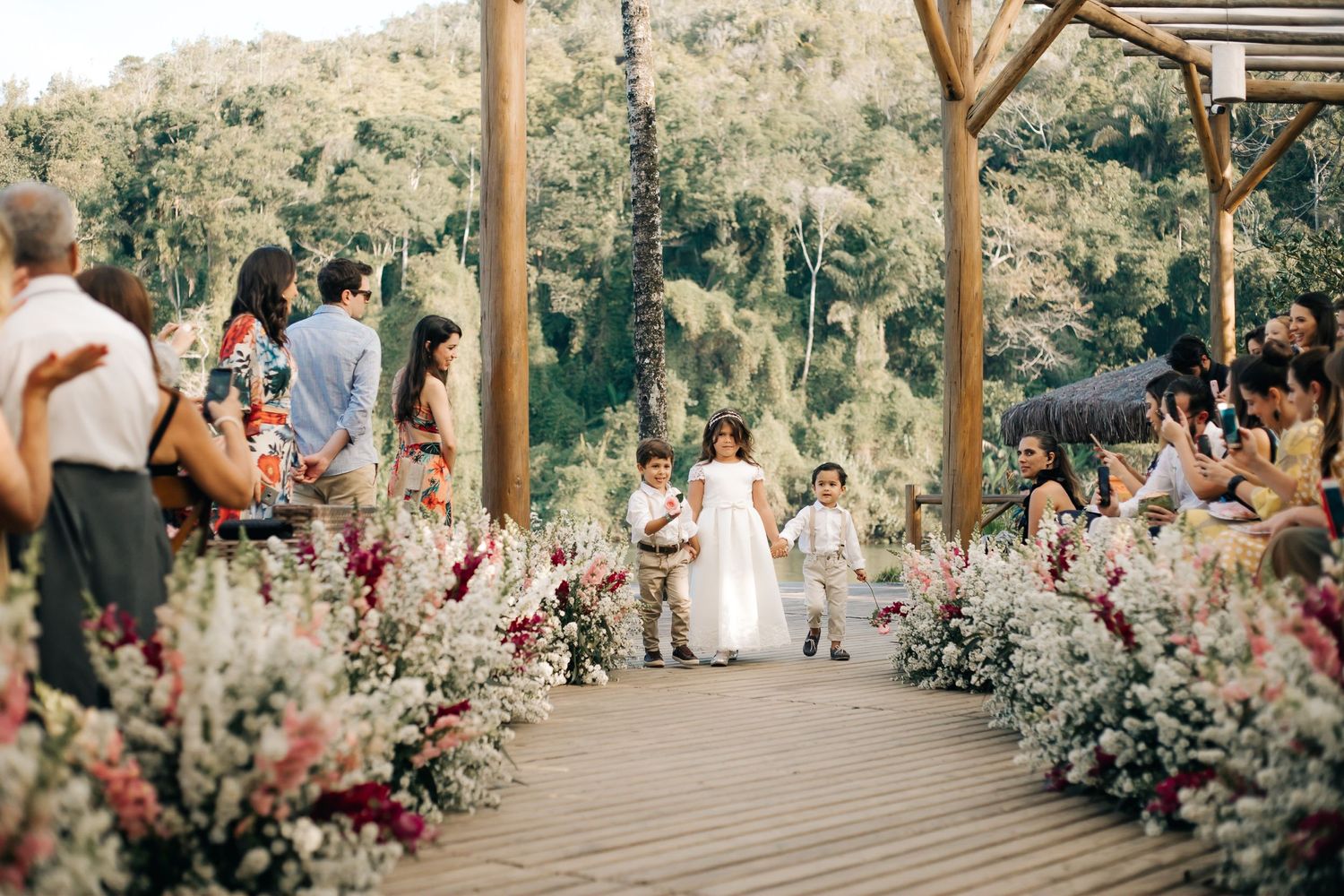 Guests seated along wooden walkway lined with pink and white floral arrangements for outdoor ceremony.