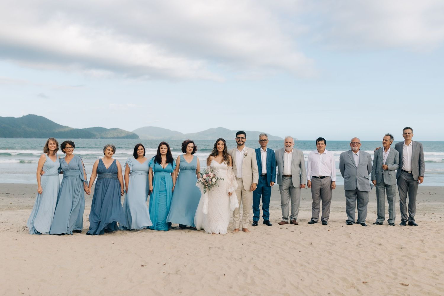 Large wedding party in coordinated blue and white outfits lined up along scenic beach.