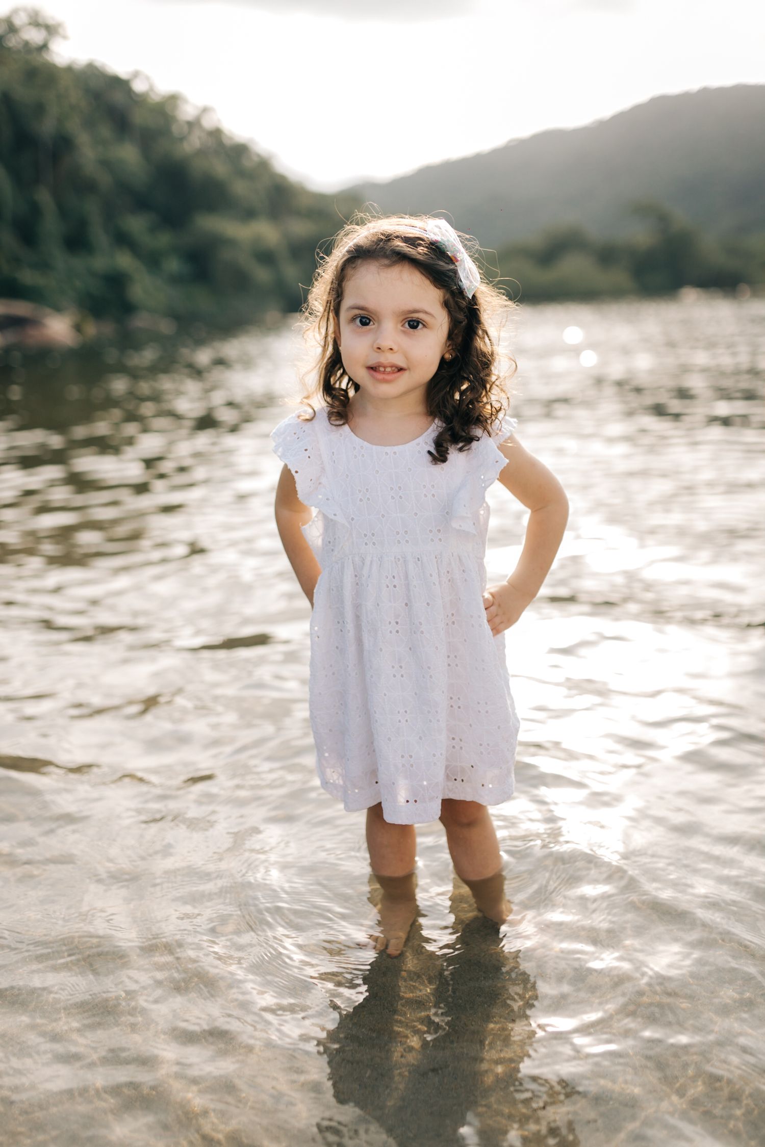 A young child stands in shallow water wearing a white dress against a mountain backdrop.