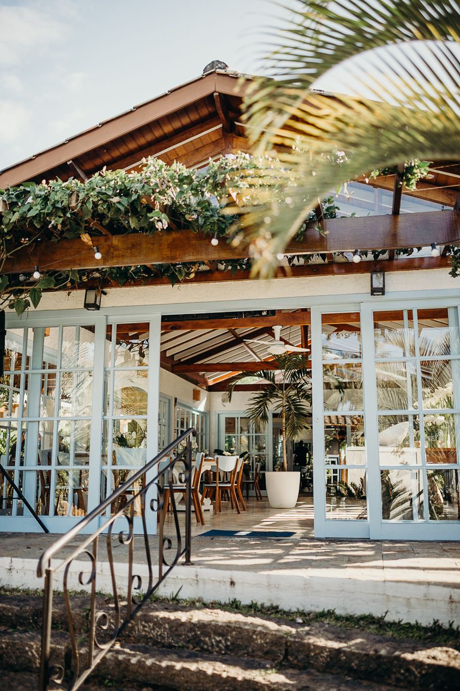 Tropical restaurant entrance with wooden beams, glass doors, and lush greenery decorations.