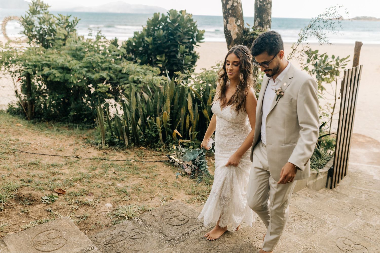 Destination wedding couple walking along tropical coastal path in elegant attire.