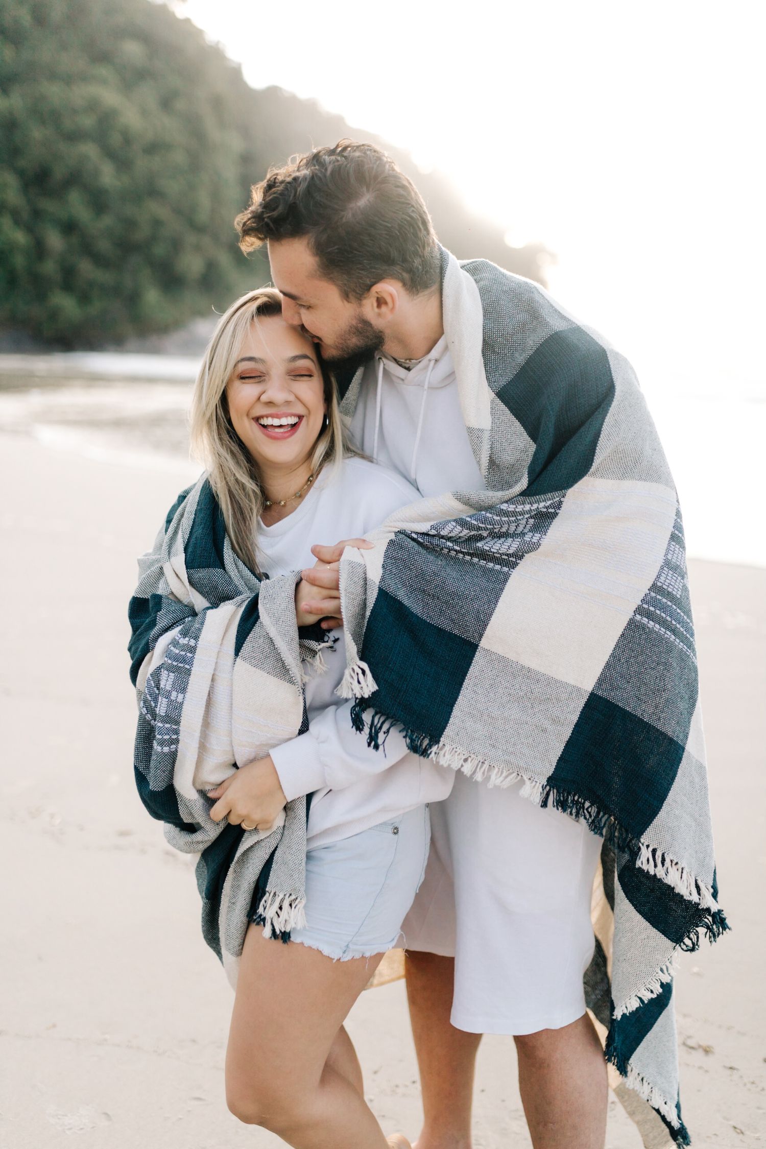 A couple shares a joyful moment wrapped in a navy plaid blanket on a misty beach.