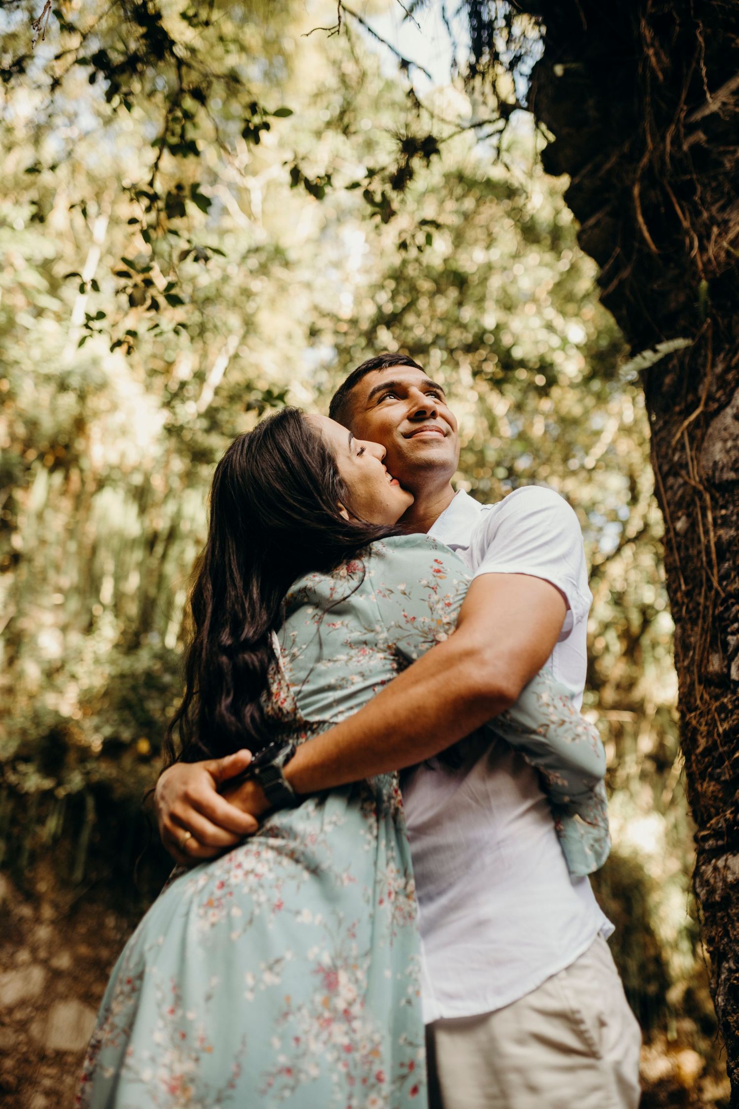 A joyful couple shares a laughing embrace in dappled sunlight under forest trees.