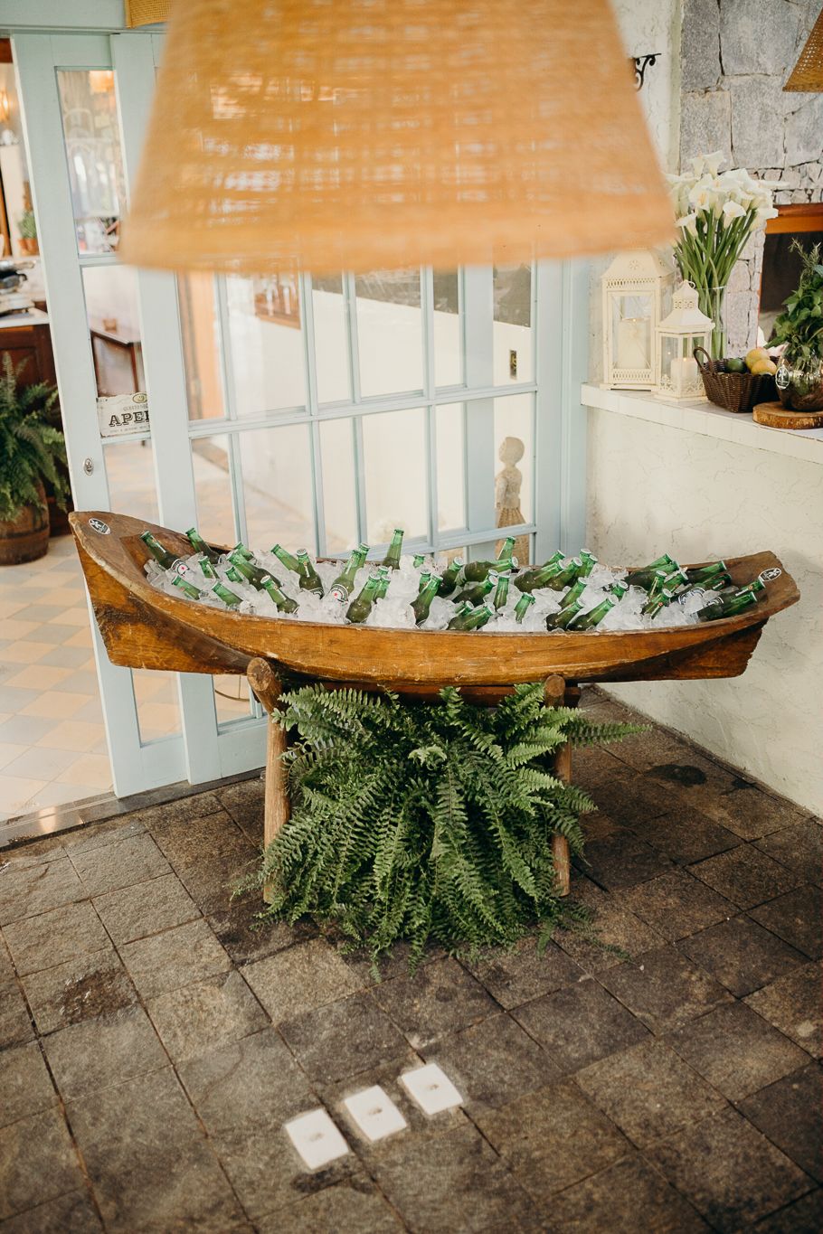 Wooden boat filled with drinks under an orange lamp shade next to a decorative fern.