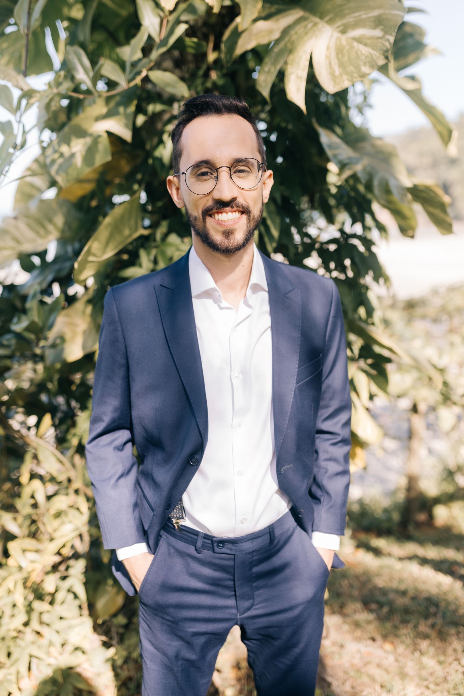 Groom in navy suit and glasses poses against tropical foliage background.