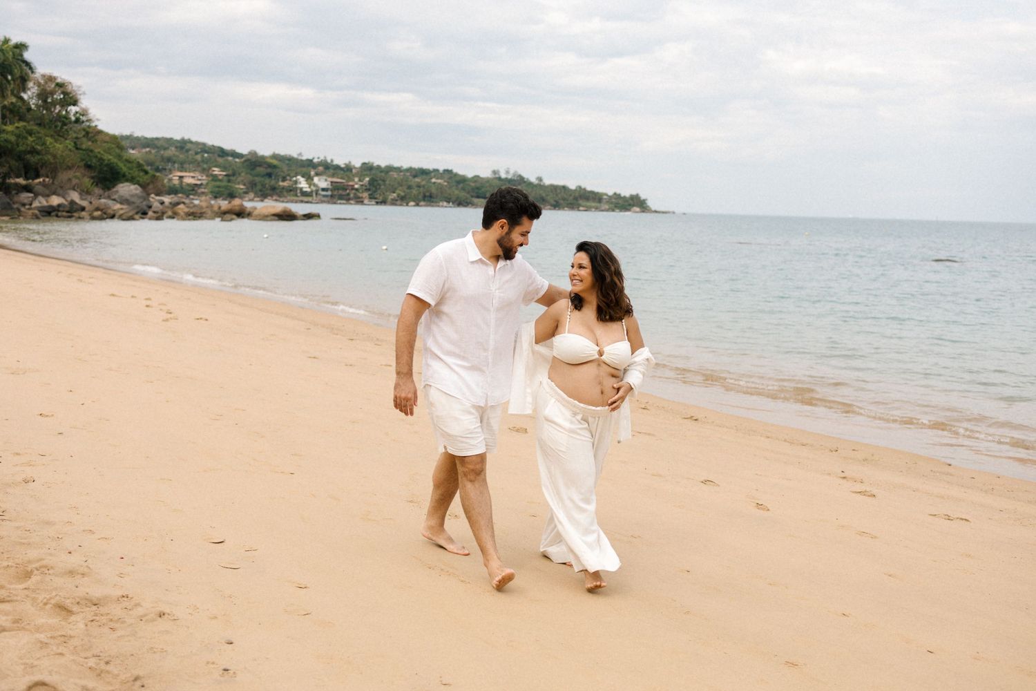 A couple in white outfits walking together on a sandy tropical beach with ocean waves in the background.