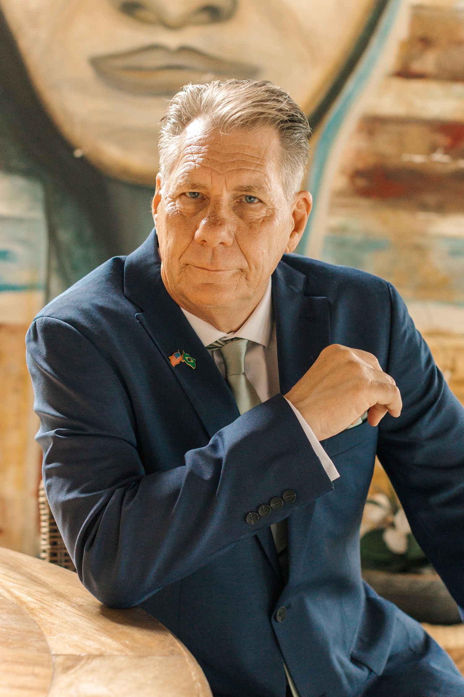 A person in a navy suit smiles warmly while sitting in a casual indoor setting.