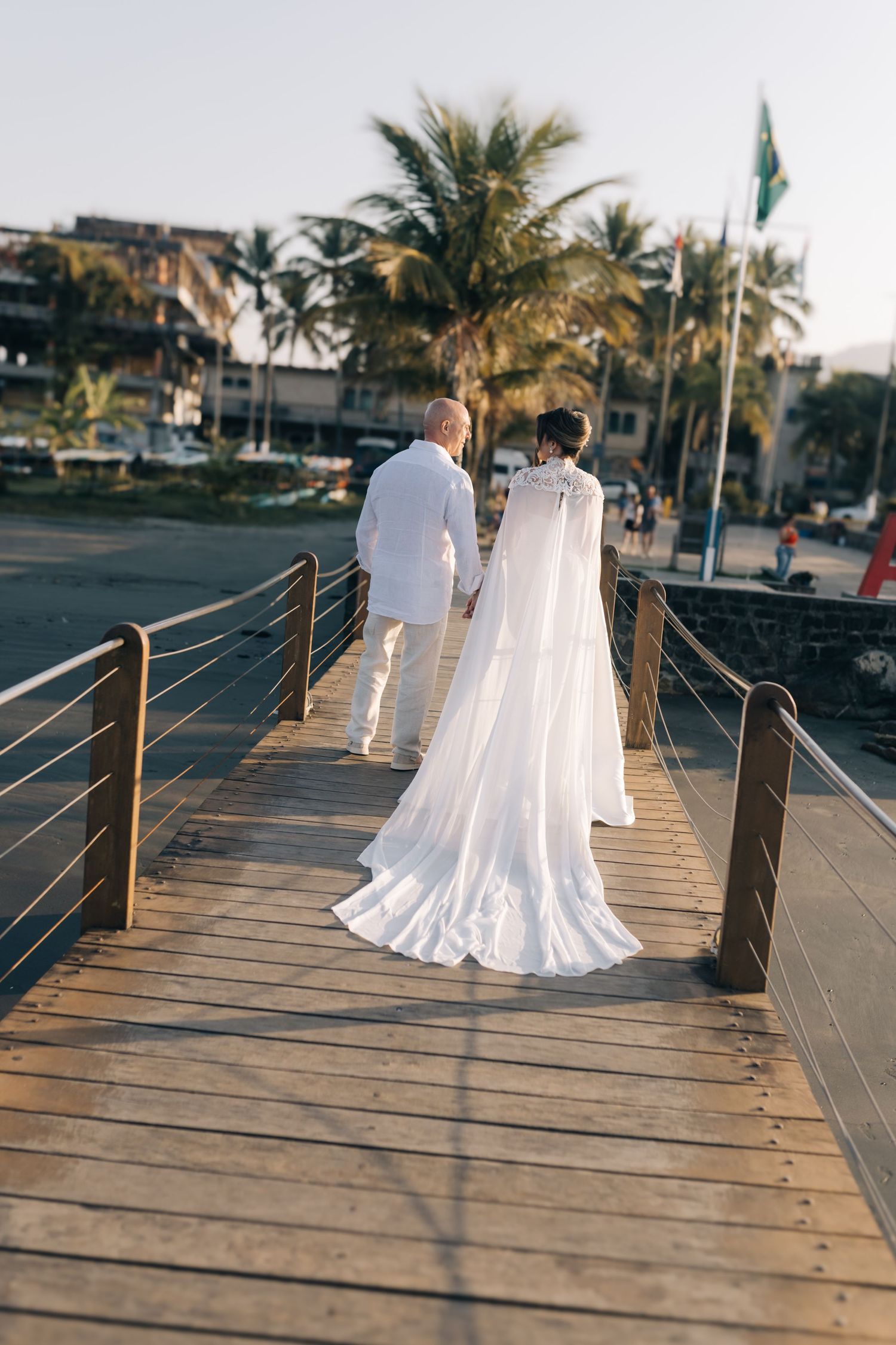 A couple in white wedding attire walks on a wooden boardwalk near palm trees during dusk.