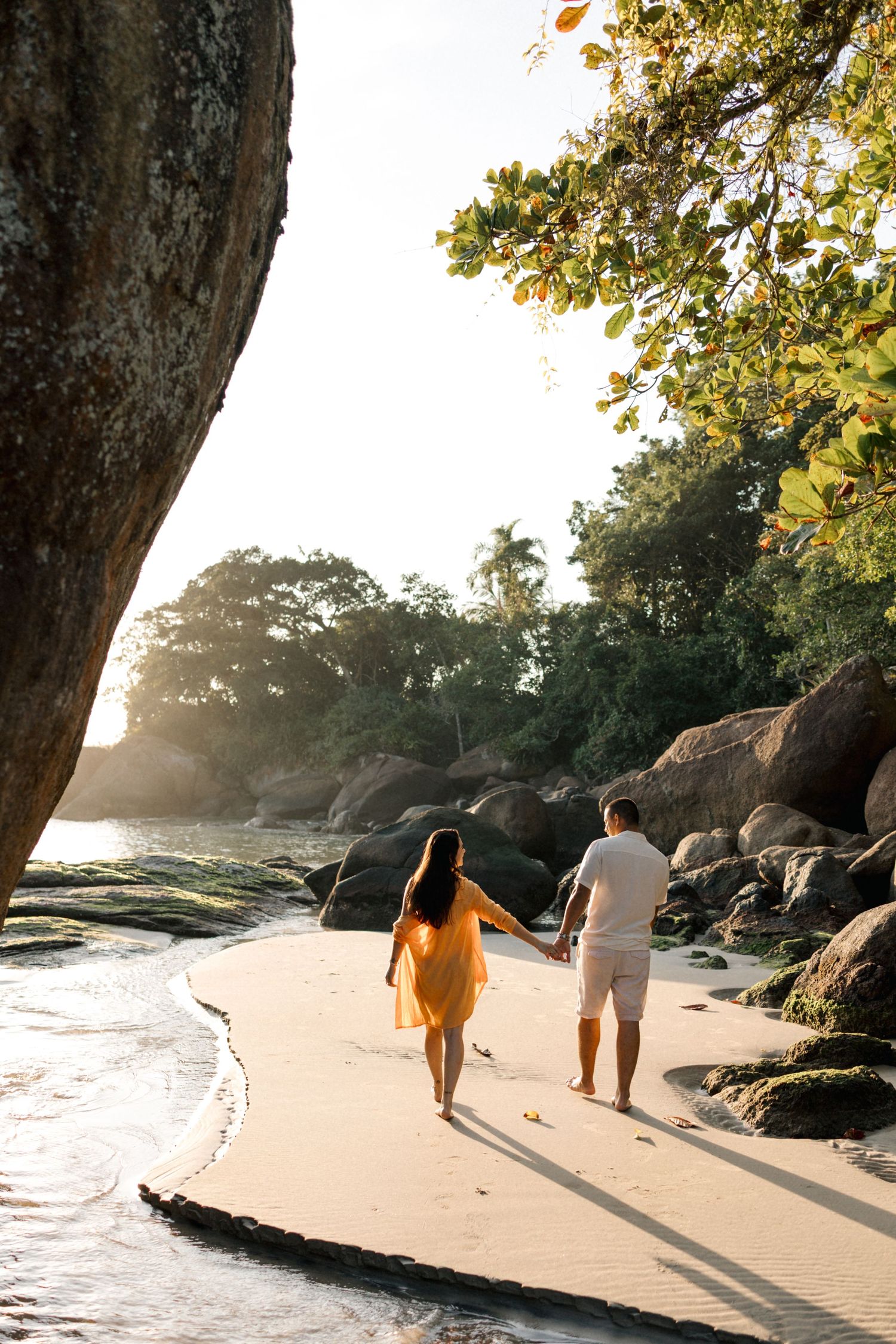 Couple walking hand in hand on beach at sunset with rocky coastline in background.