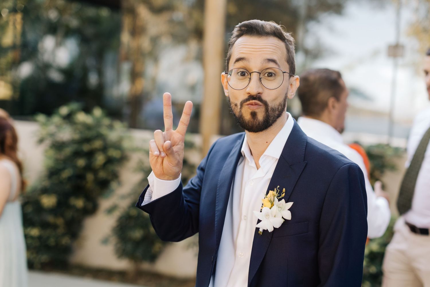 Wedding guest in navy suit and glasses making peace sign while wearing a white boutonniere at outdoor celebration.