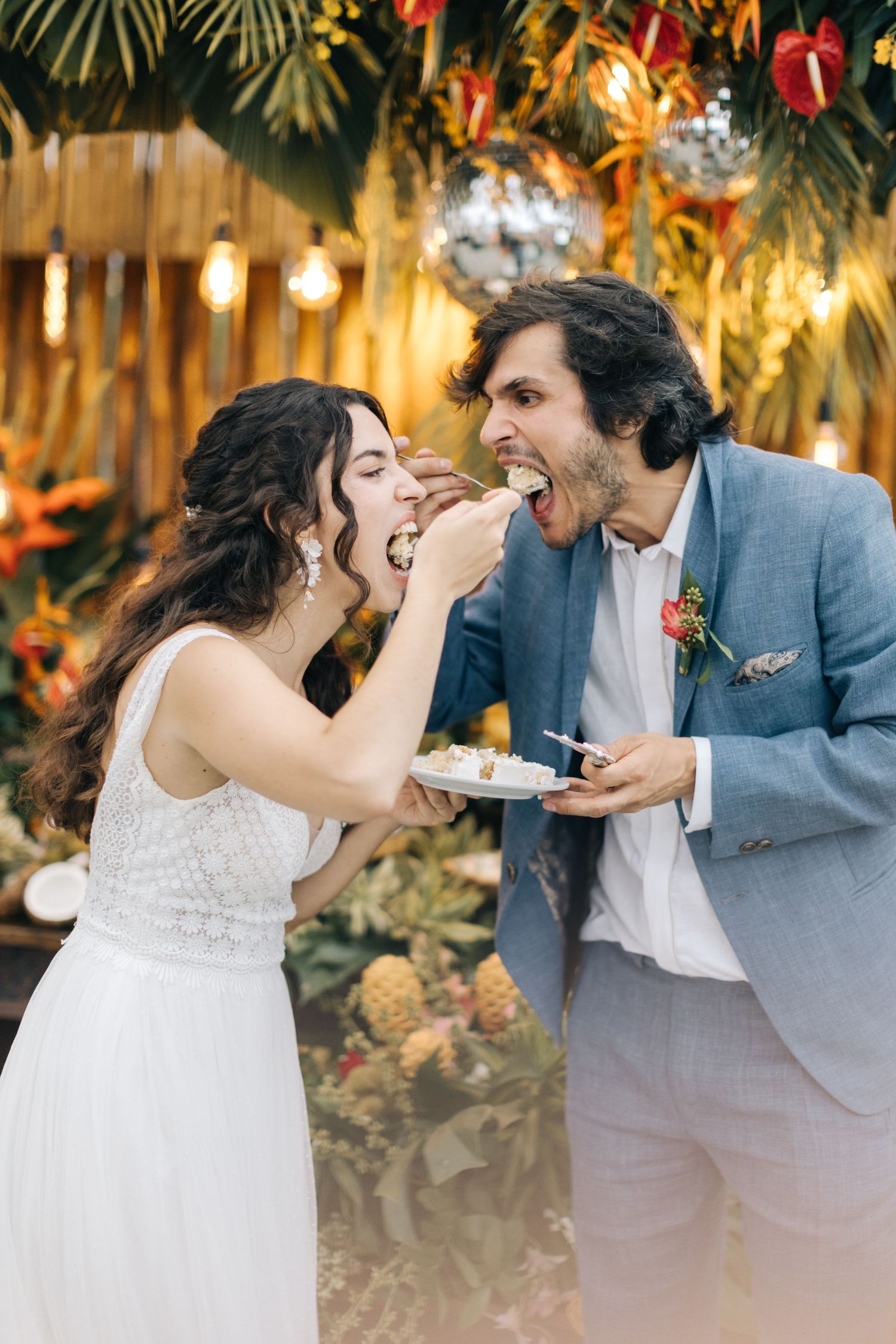 Playful couple feeding each other wedding cake under tropical decor and disco balls.