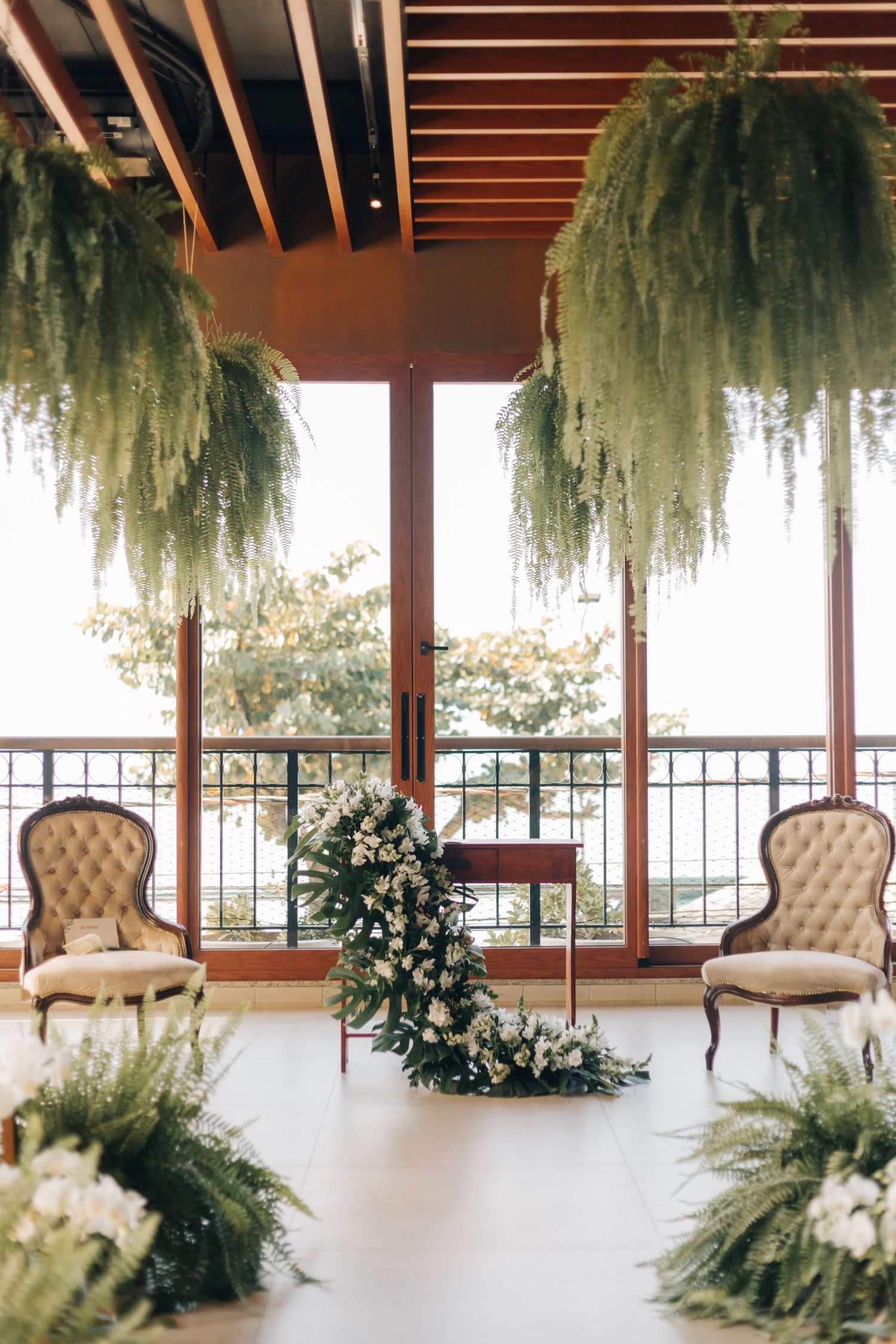 Elegant wedding ceremony setup with vintage chairs and hanging greenery in front of large windows.