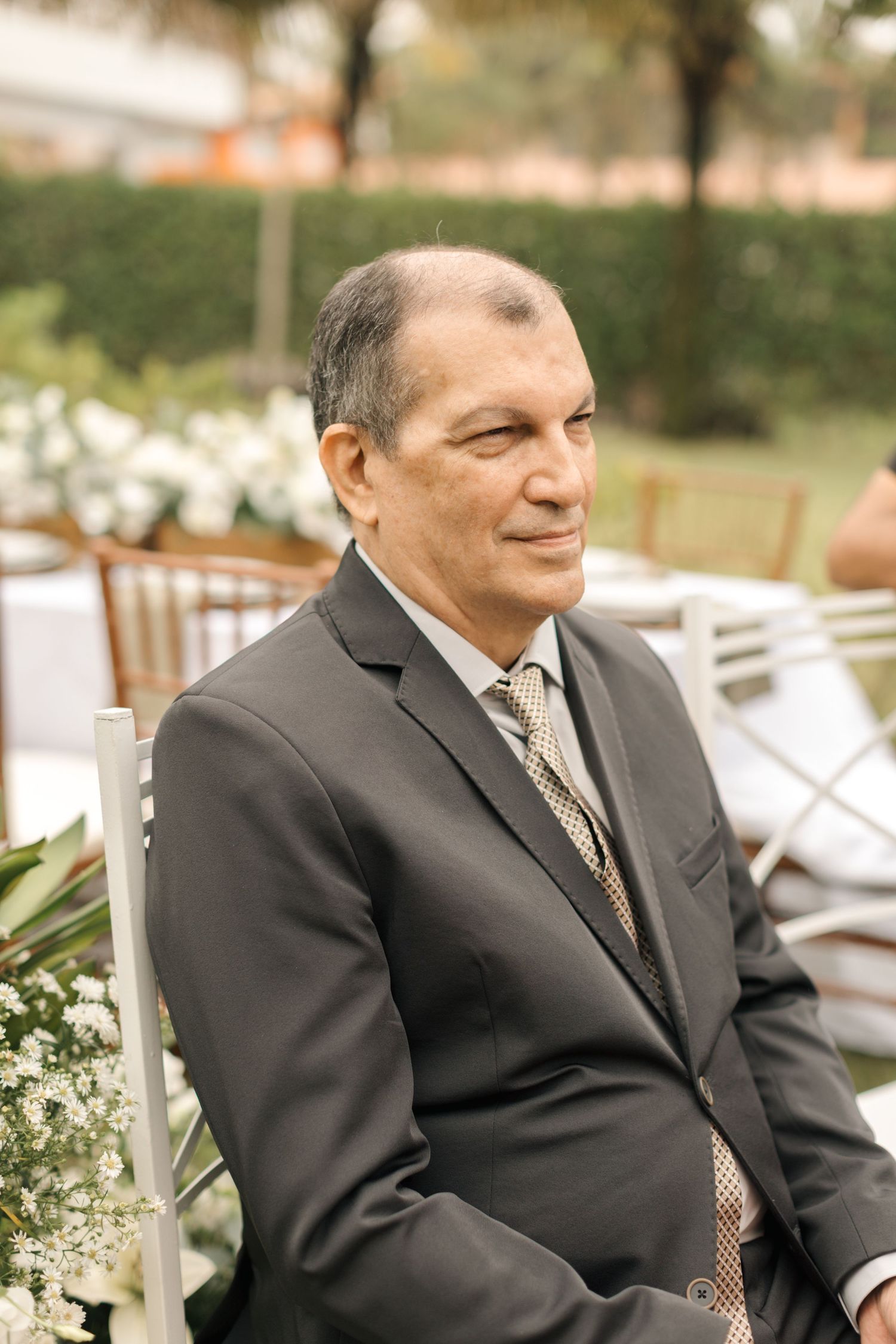 A formally dressed man in a dark suit and tie sits at an outdoor event.