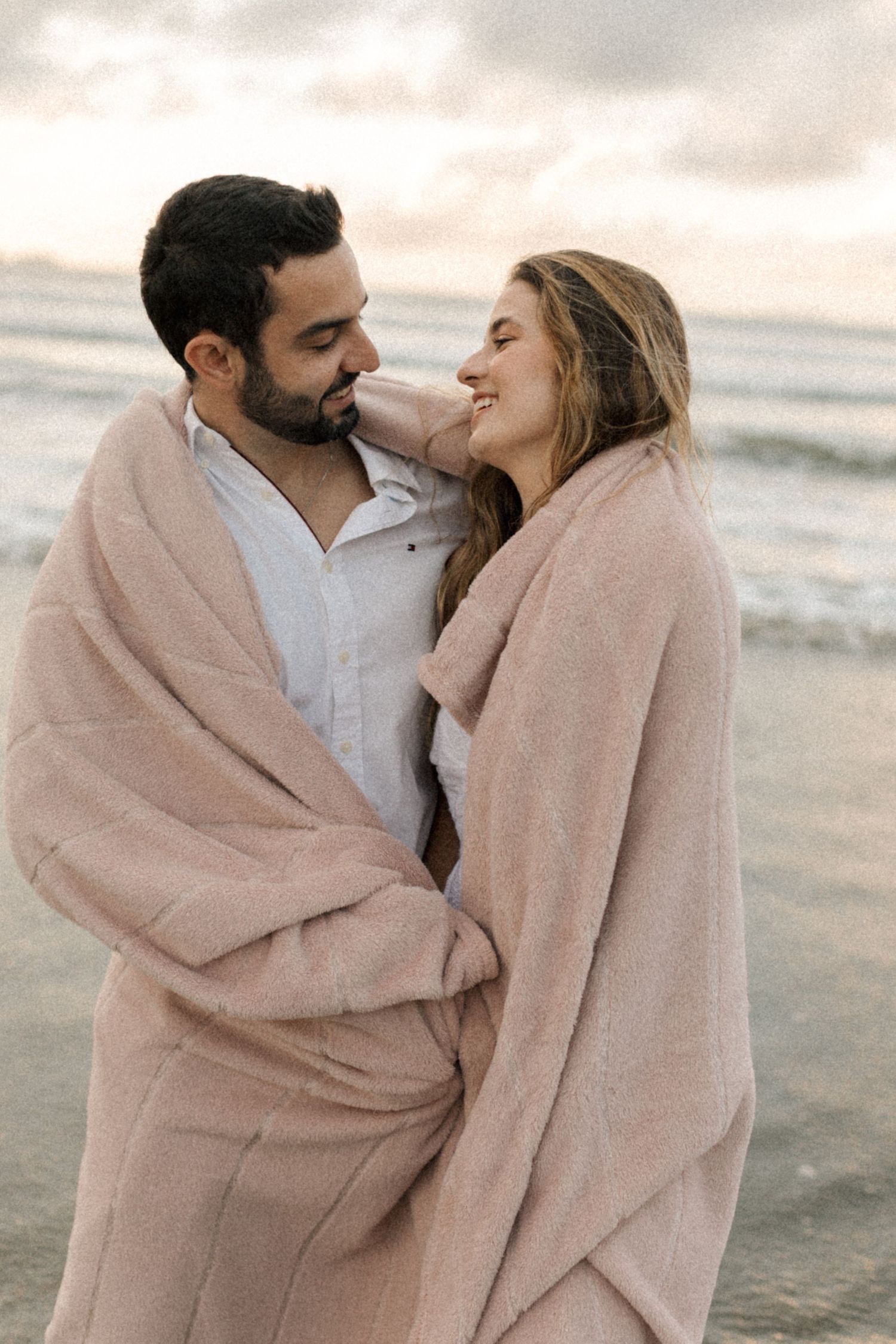 Two people embrace while sharing a soft pink blanket on a misty beach shore.
