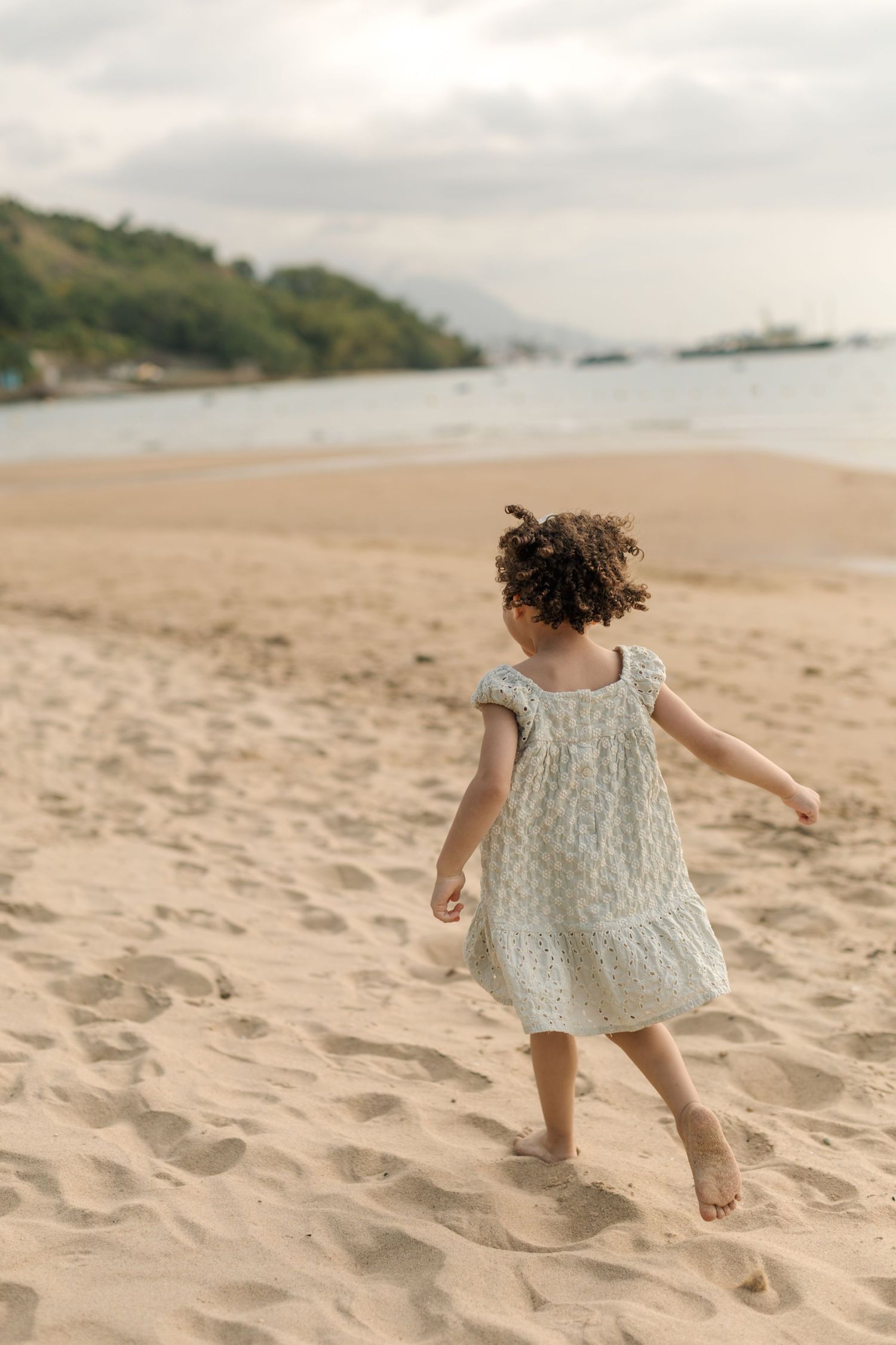 A small child in a white dress twirls on a sandy beach with boats and hills visible in the background.
