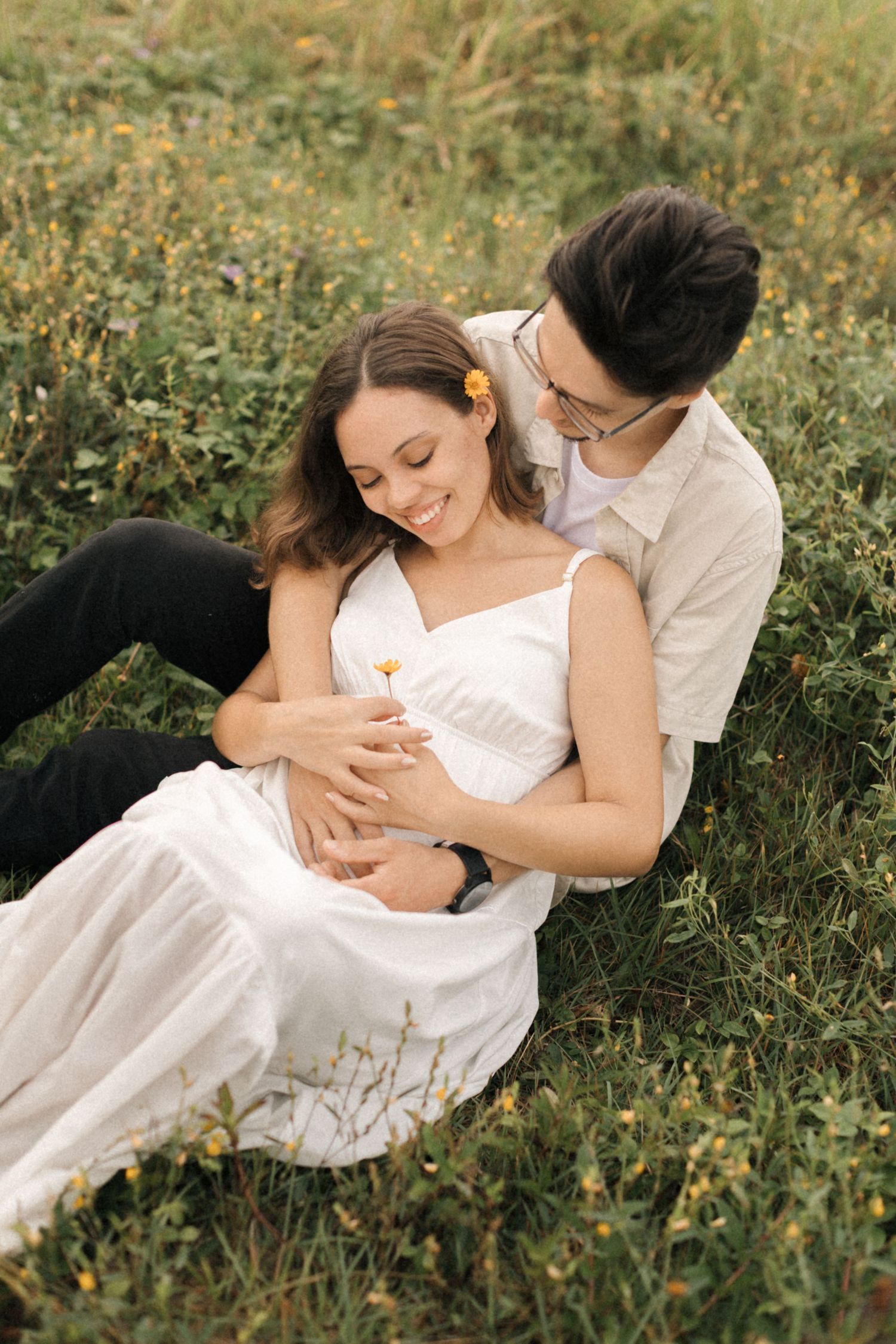 A loving couple embraces while lying in a sunny meadow filled with wildflowers and green grass.