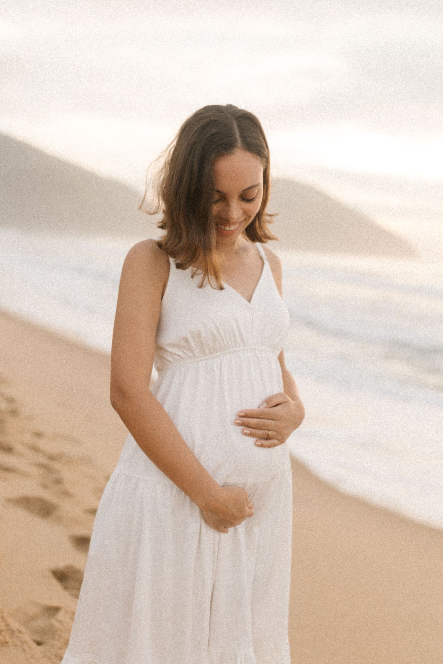 Woman in a flowing white dress cradles her baby bump while standing on a beach at sunset.