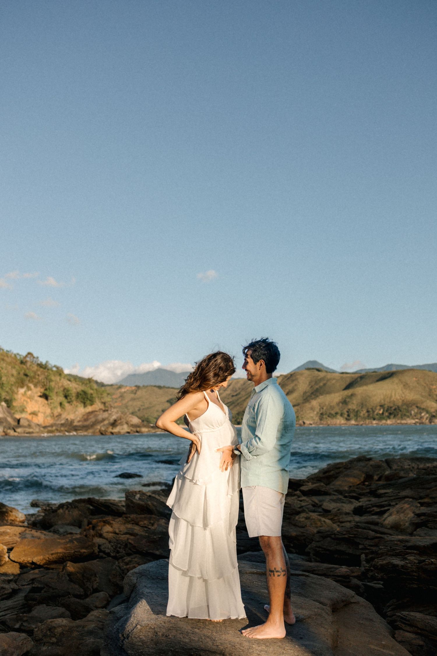 A couple poses on rocky coastal terrain with mountains and ocean views.