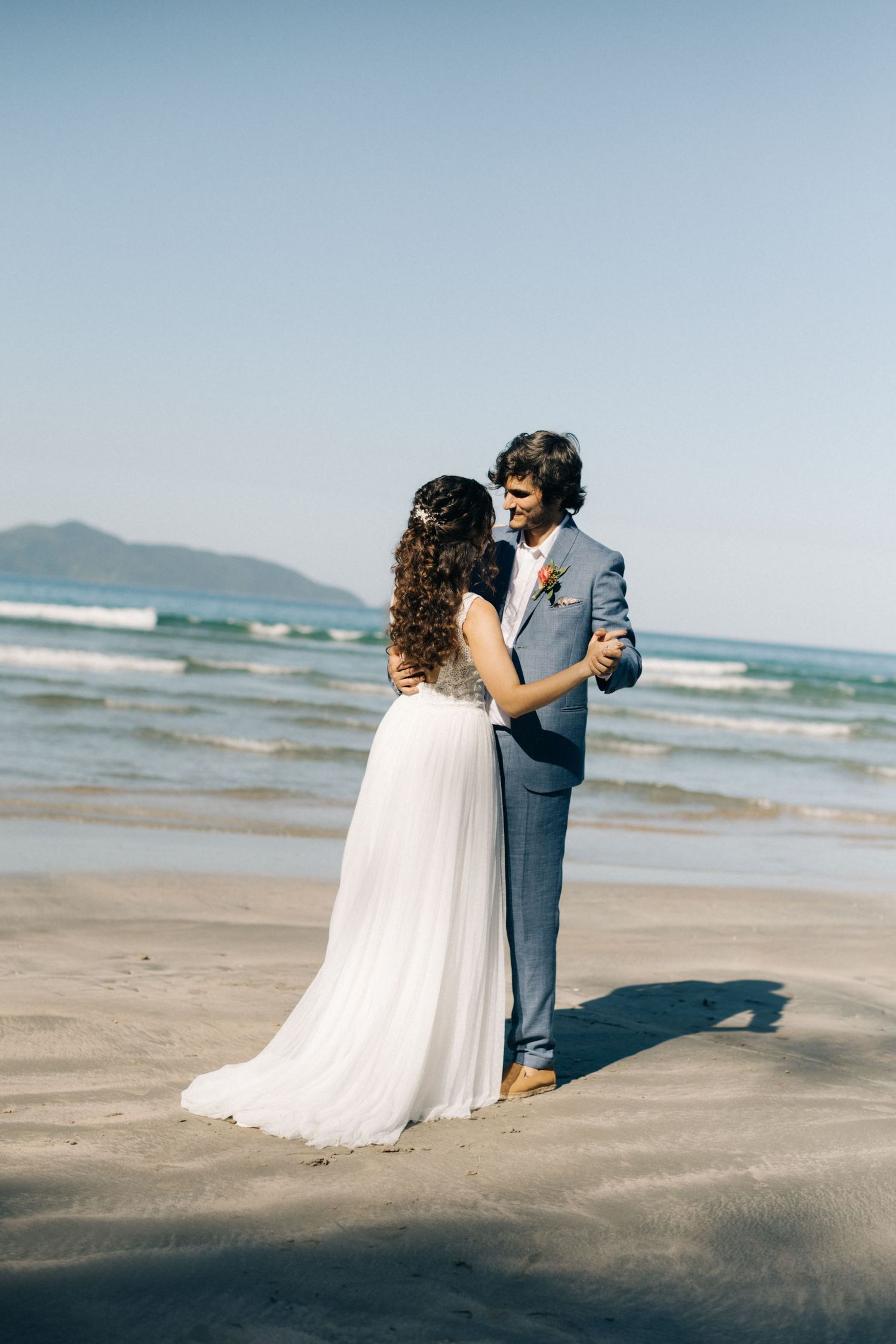 Couple dances on oceanfront beach with waves and mountains in background.