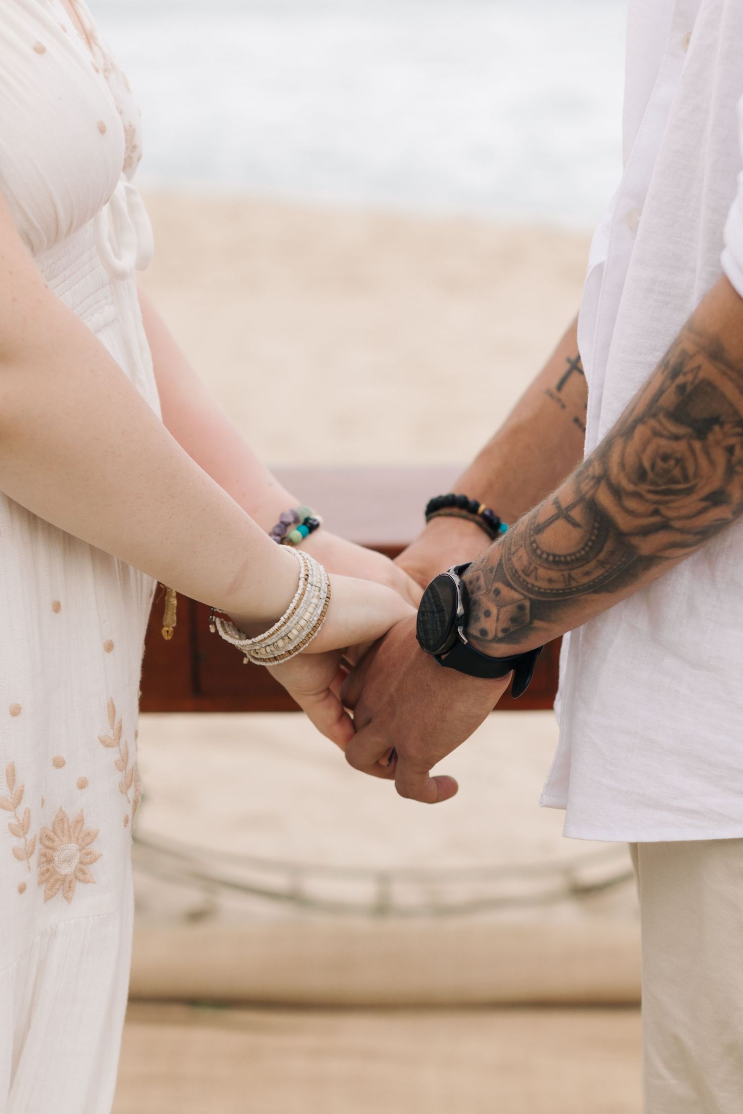 Close-up of tattooed hands clasping together with bracelets and jewelry visible against a beach backdrop.