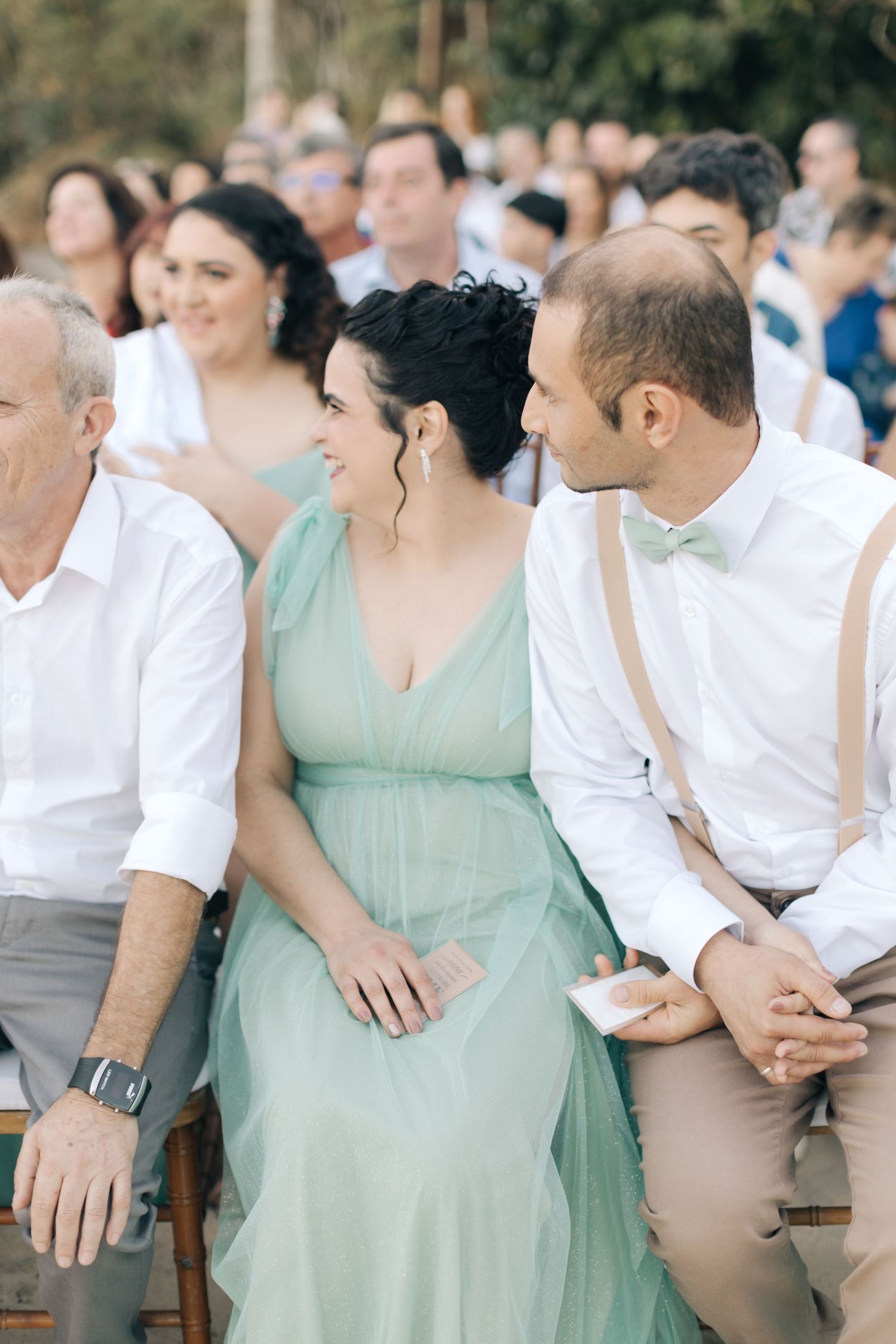 Wedding guests in formal attire seated on wooden benches at an outdoor ceremony.