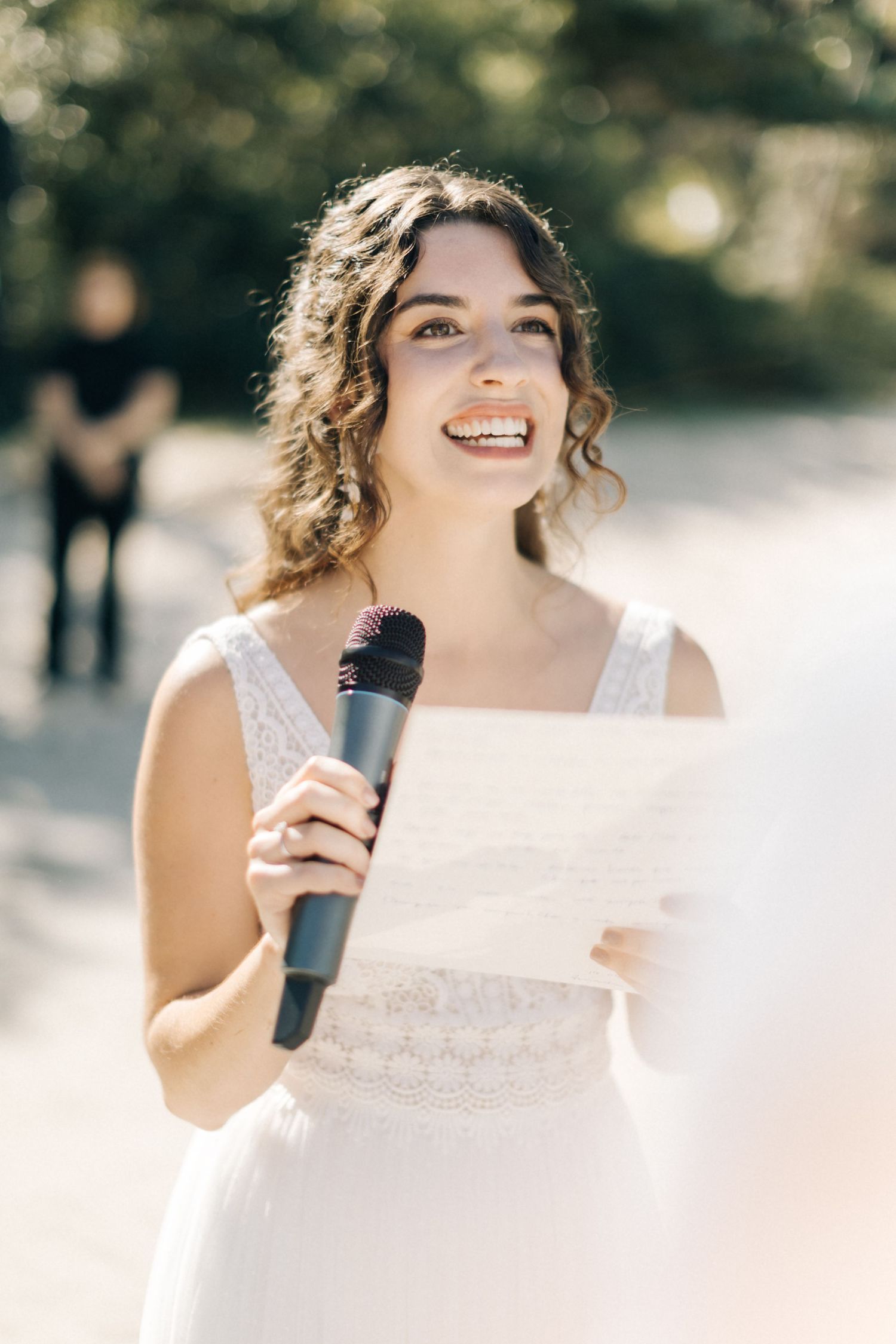 Bride holding microphone and paper while speaking outdoors at wedding ceremony.