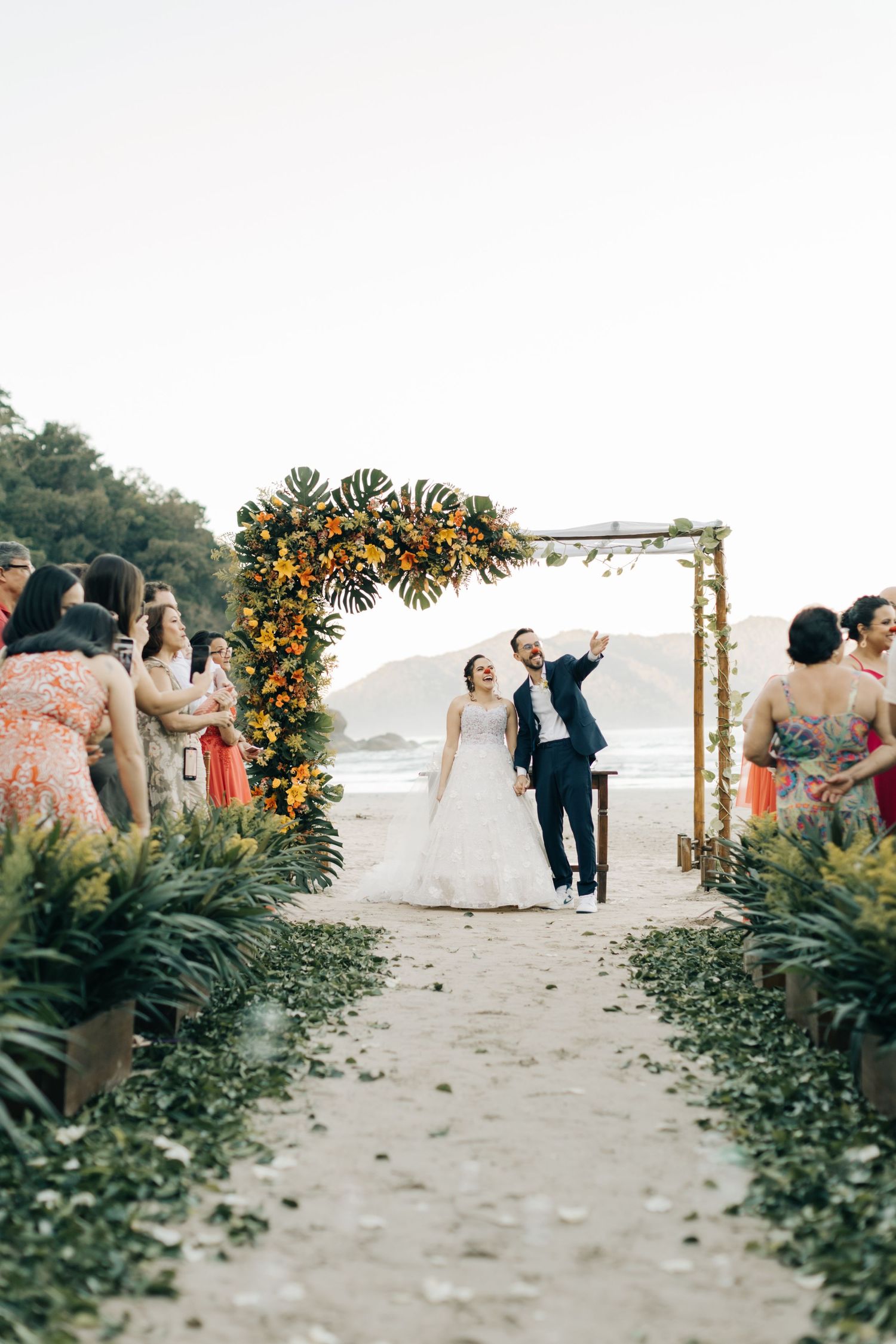 Wedding couple celebrates under floral arch while guests line pathway on beach.