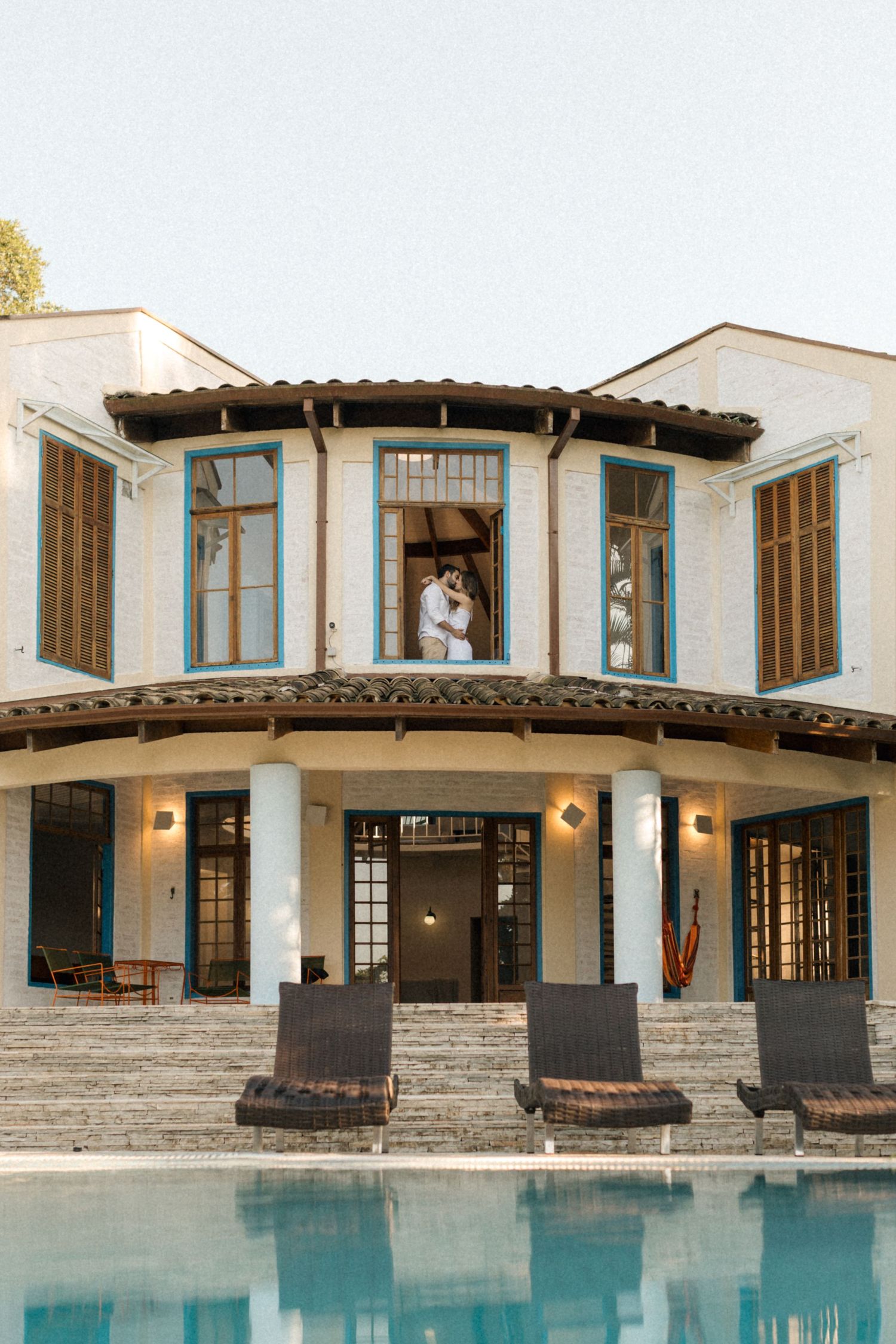 Elegant two-story villa with a curved balcony and French doors overlooking a serene pool.