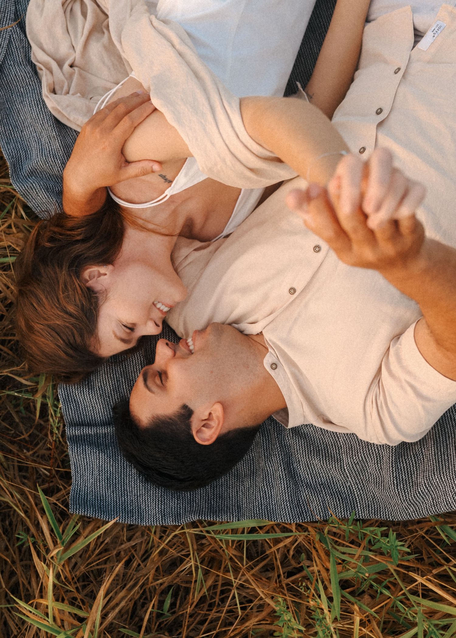 Two people in light colored clothing lie close together on a blue blanket outdoors.