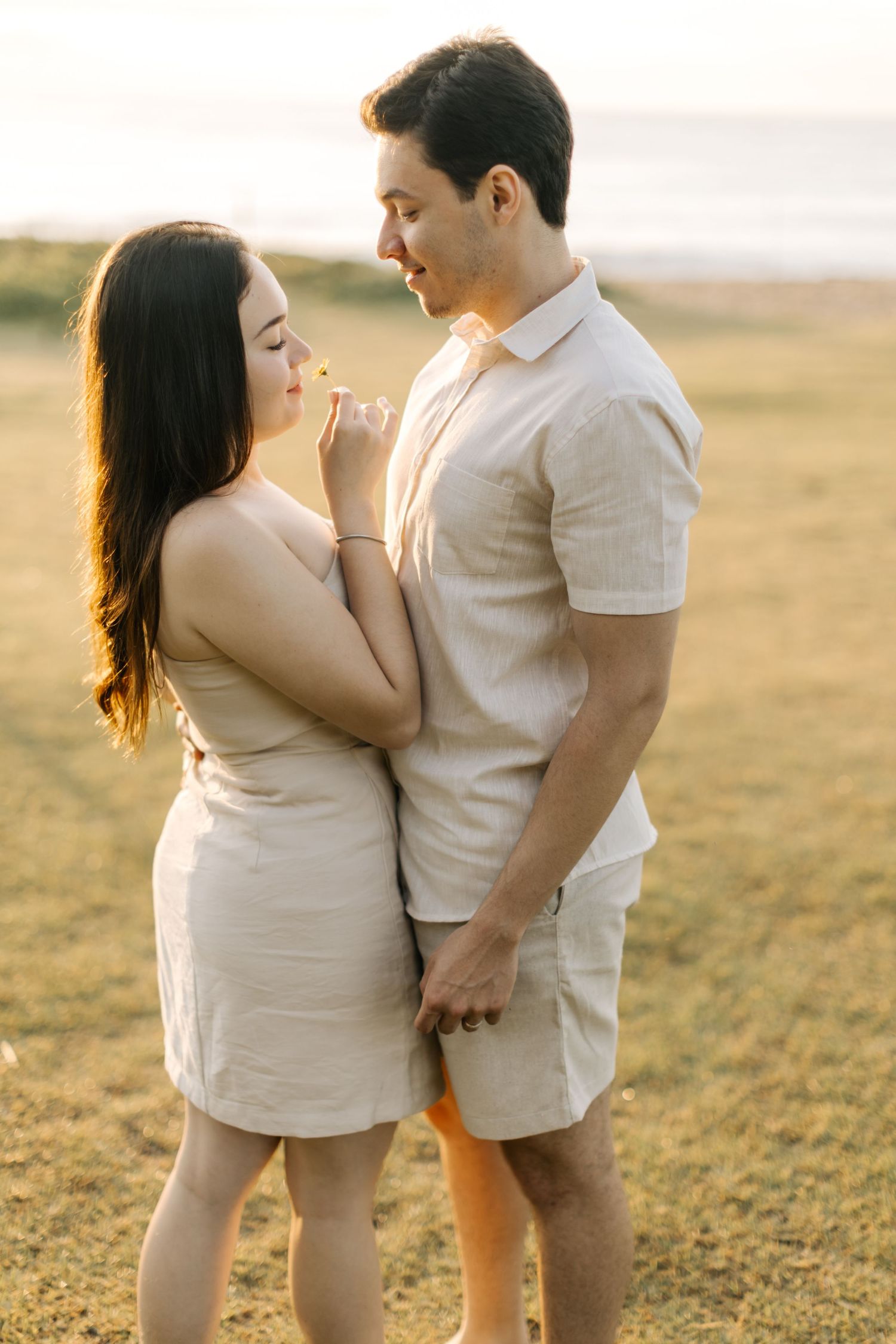 Romantic couple in matching cream outfits shares an intimate moment on a hilltop at sunset.