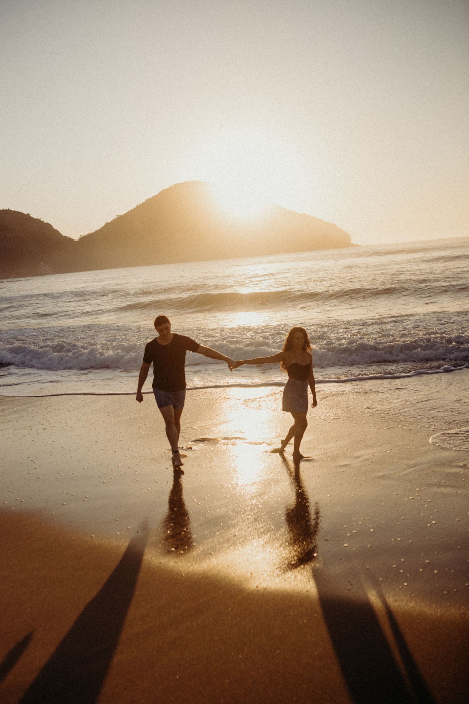 Silhouettes of a couple holding hands while walking on a beach at sunset with mountains in the background.