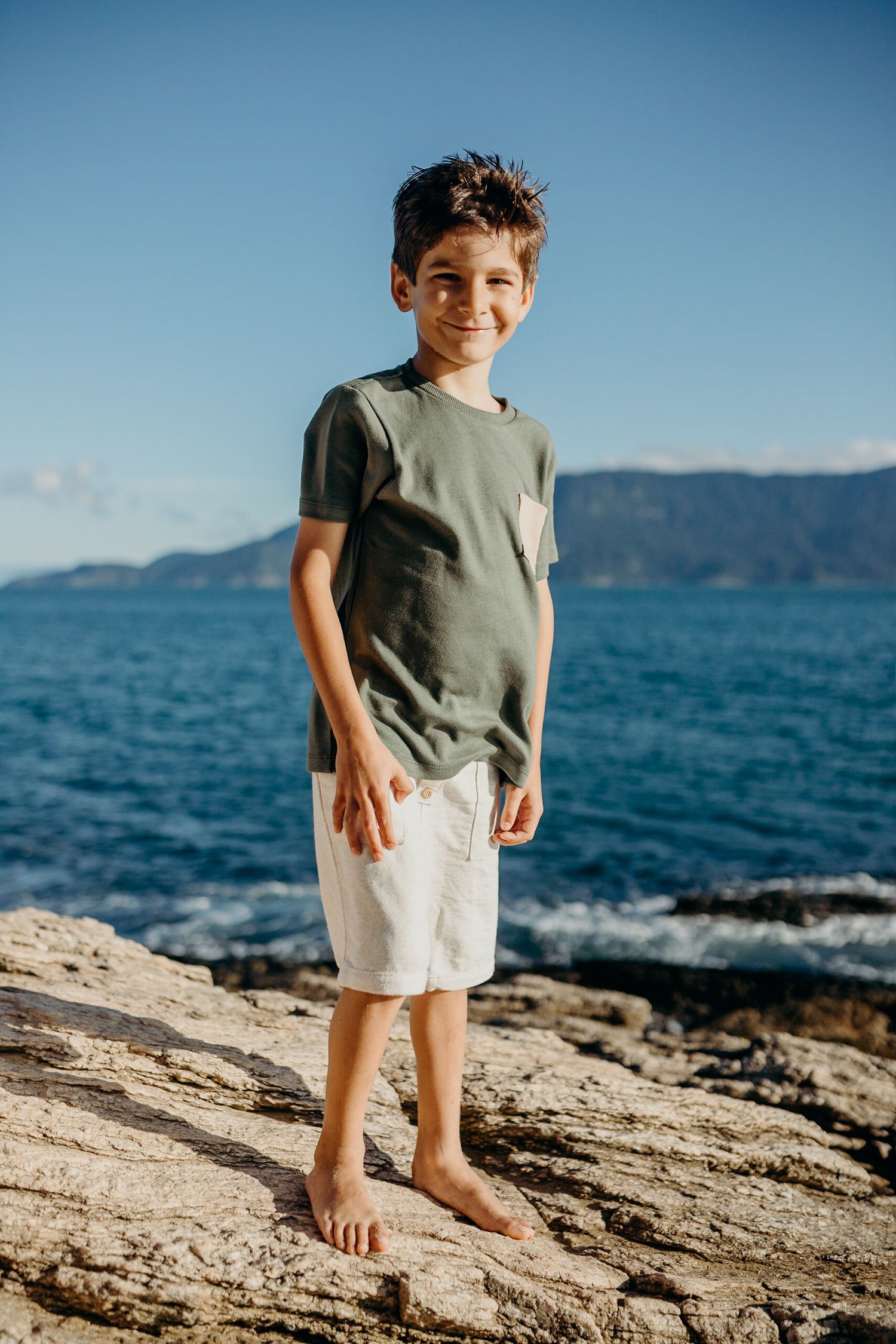 Child in gray t-shirt and white shorts stands on rocky shore with ocean view behind.