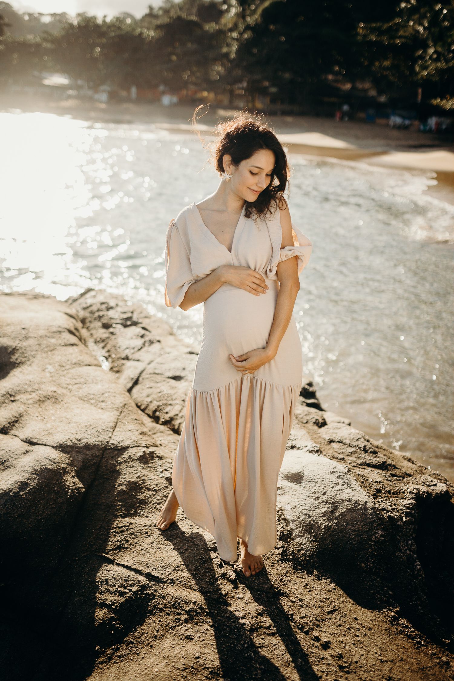 Maternity portrait on rocky beach at golden hour showing pregnant person in flowing dress.