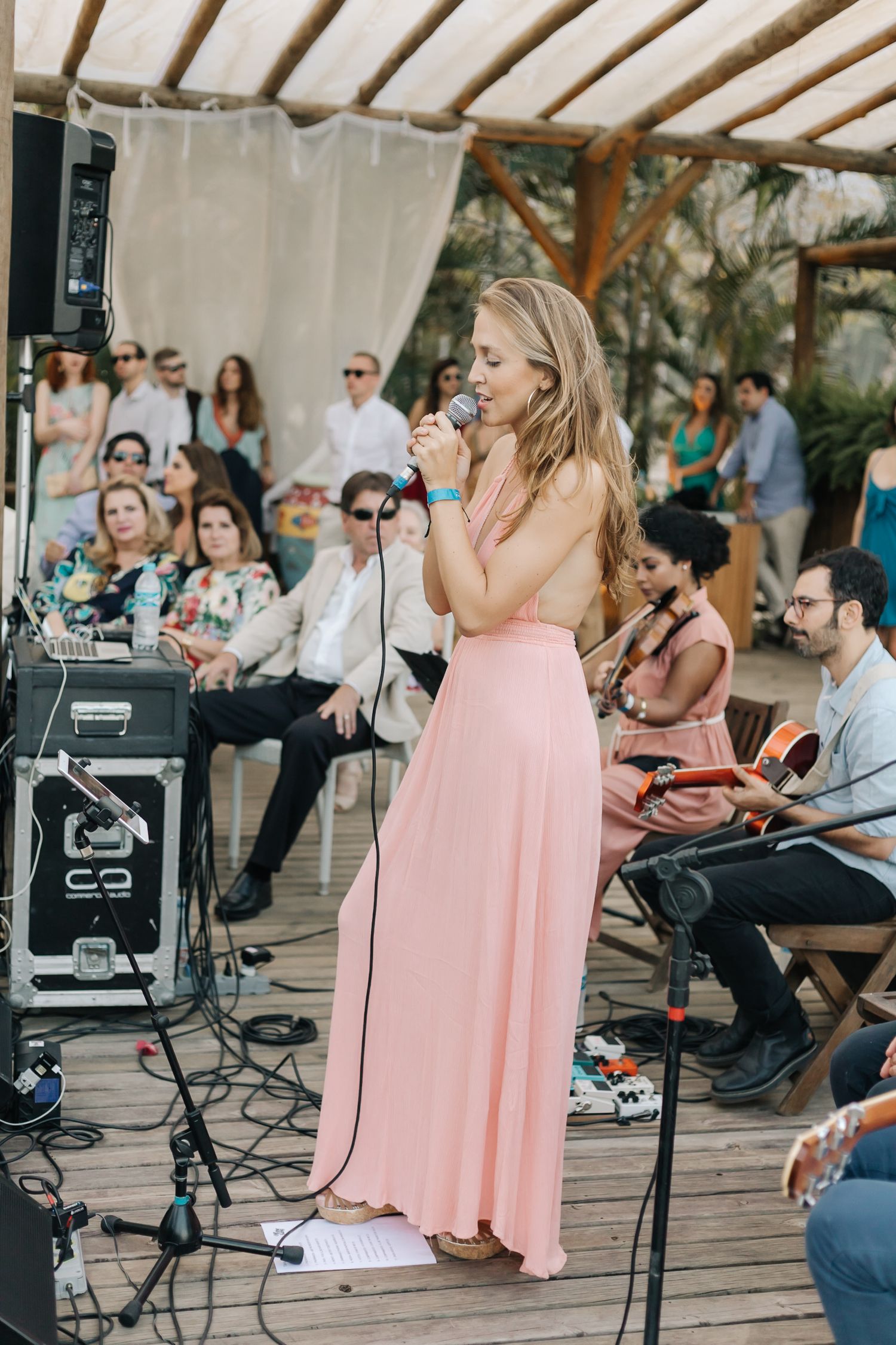 A performer in a pink gown singing at an outdoor event with musical equipment and audience visible.