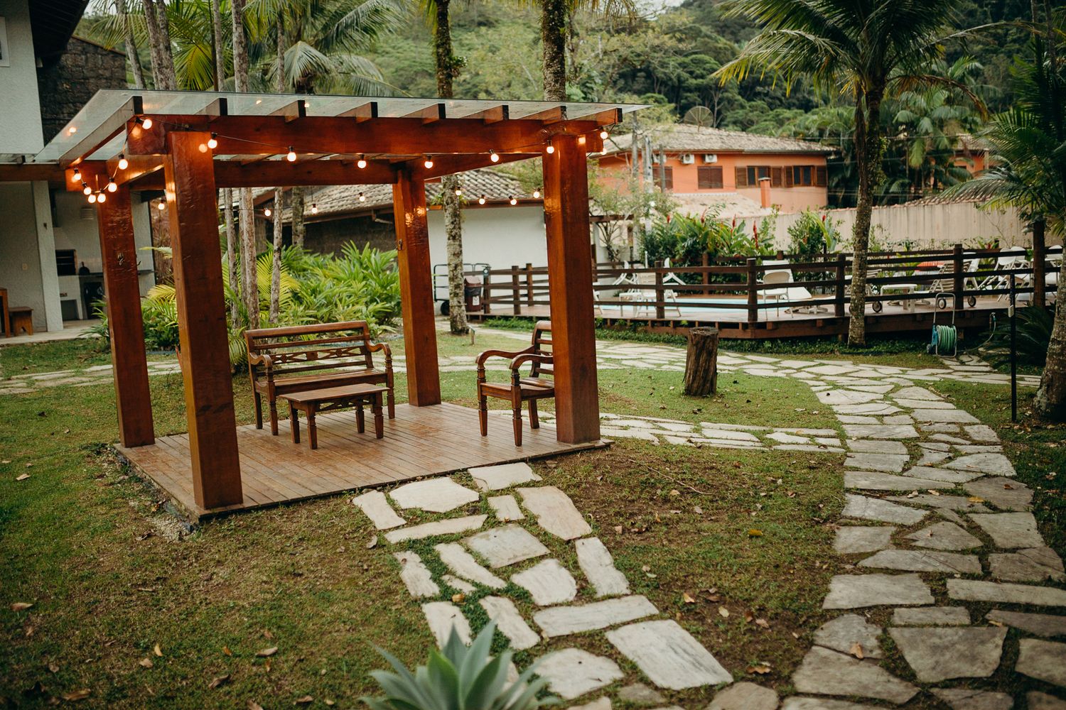 Wooden pergola with benches and stone pathway in tropical garden setting.