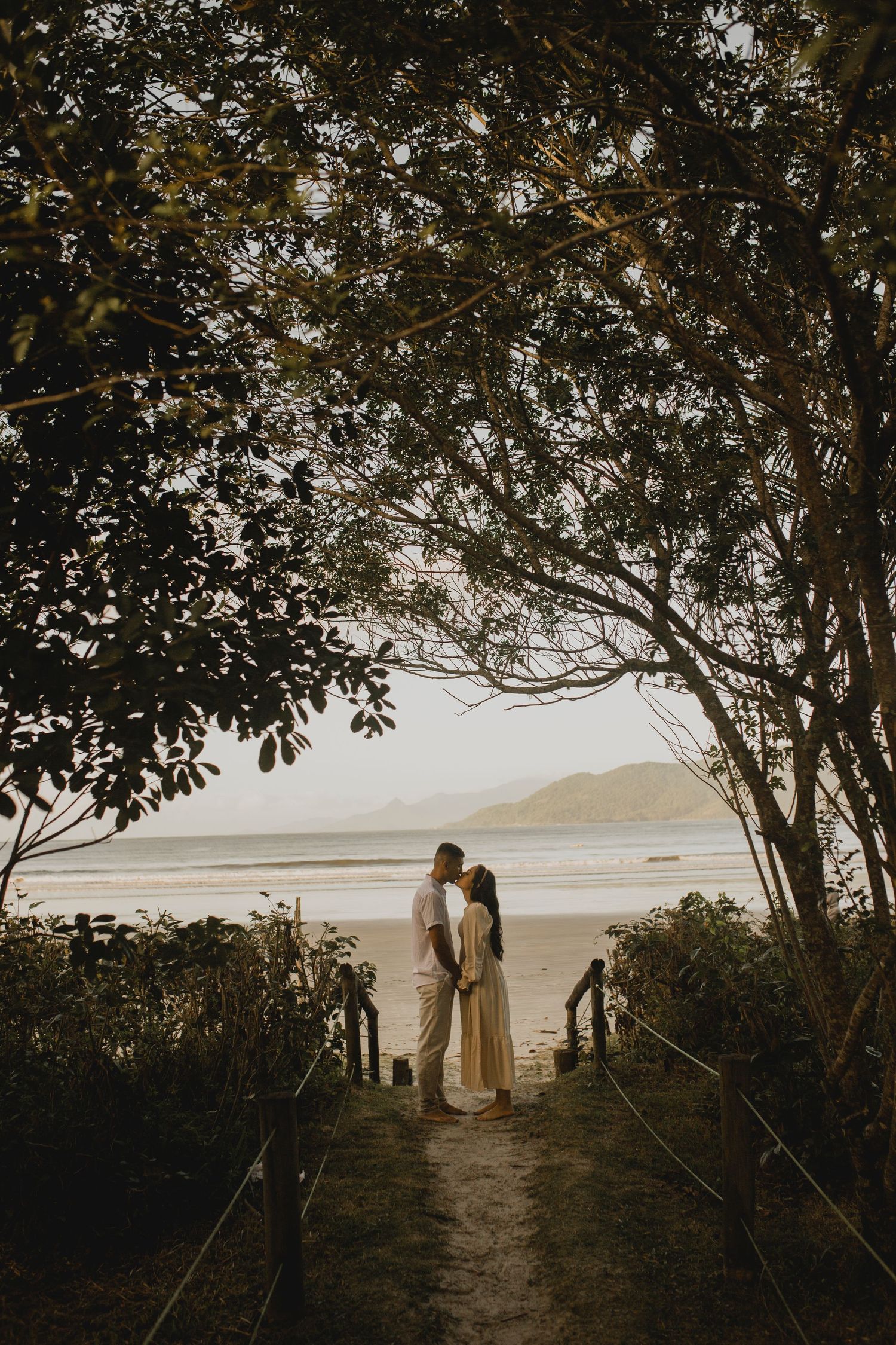 Silhouetted couple standing on pathway overlooking ocean at sunset through tree canopy.