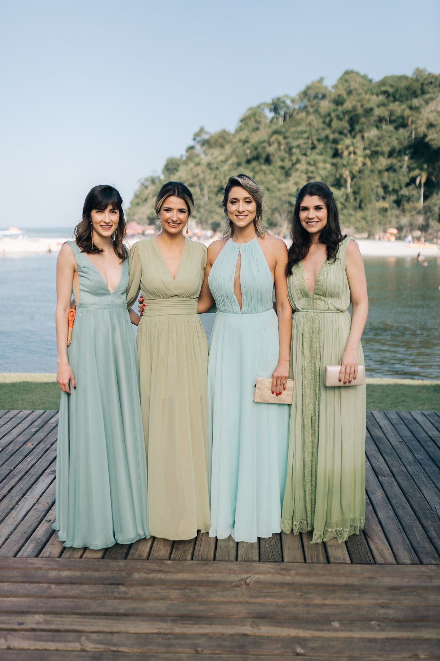 Bridesmaids wearing coordinated mint green and sage dresses pose on a wooden deck by the water.