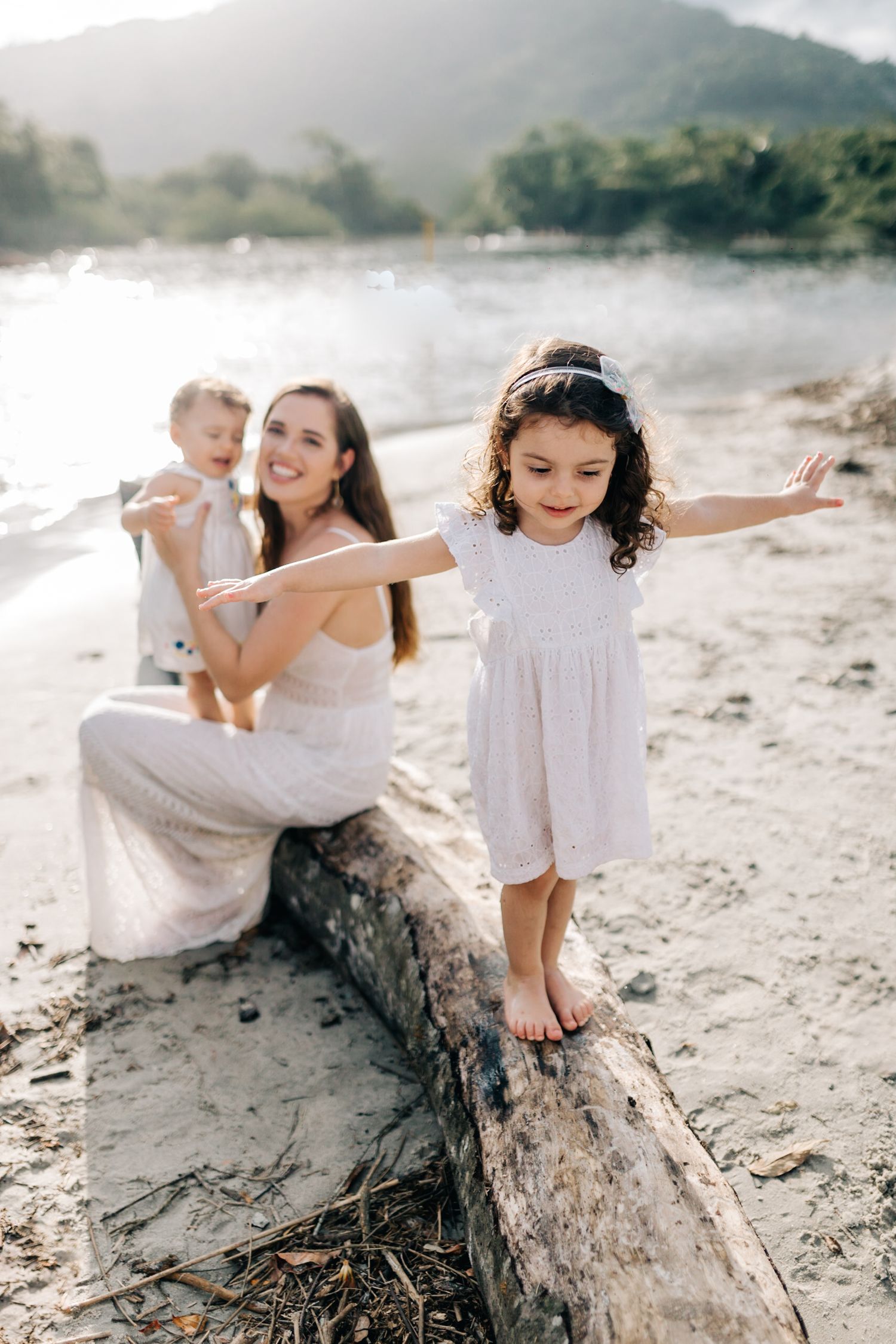 Young children balance on a driftwood log at the beach during golden hour.