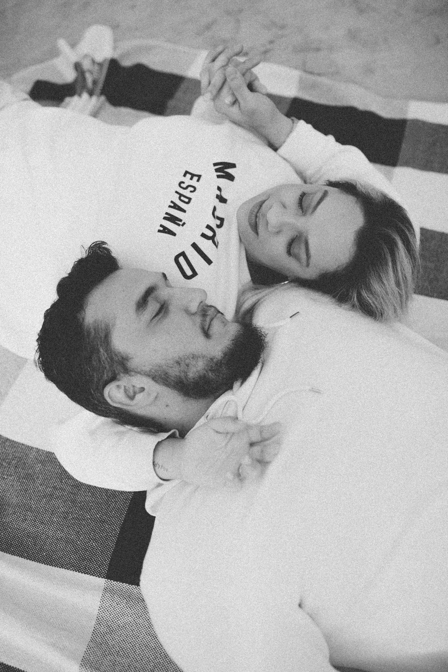 Couple in white shirts lying close together holding hands on plaid blanket.