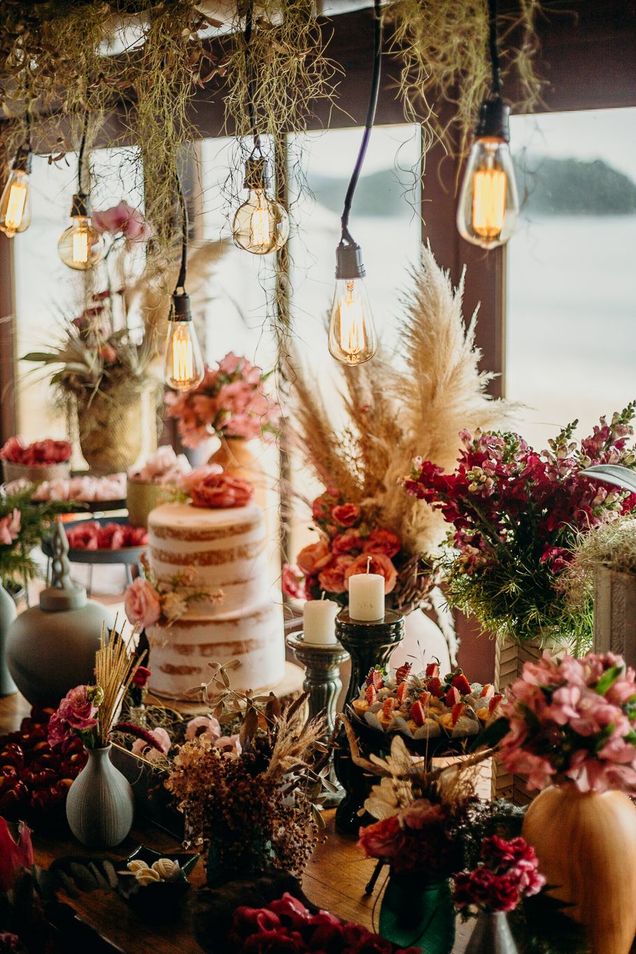 Rustic wedding dessert table with naked cake, dried flowers, and hanging Edison bulbs creating a romantic atmosphere.