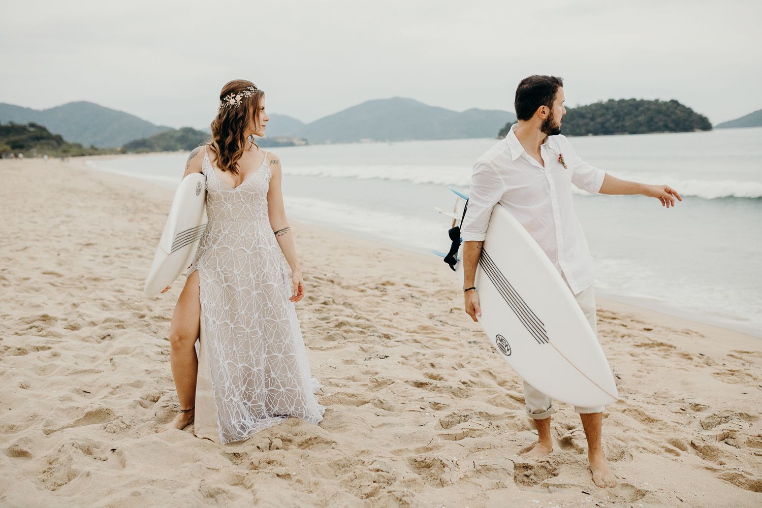 Couple in wedding attire walking on beach with surfboard and mountains in background.