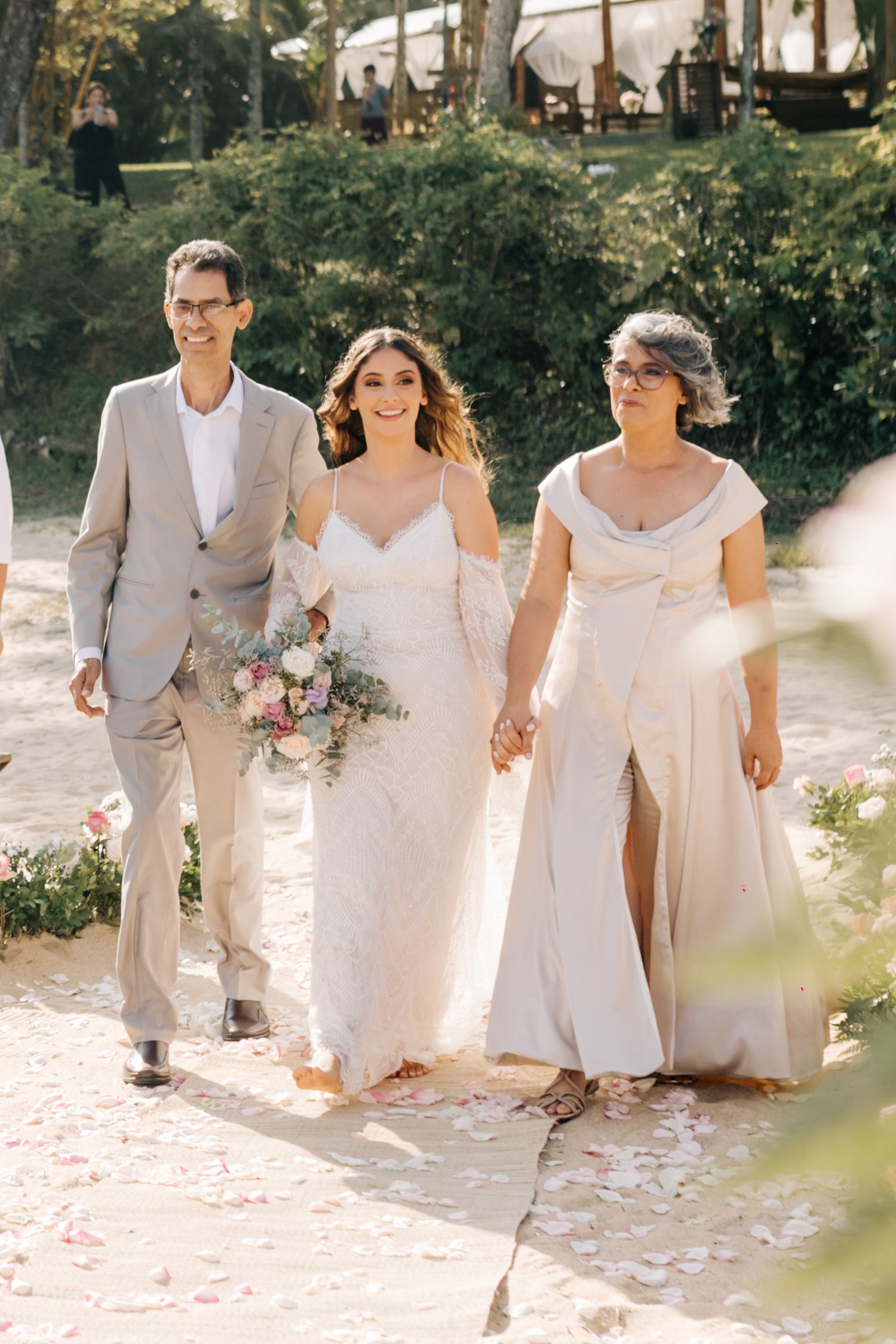 Three people walk down a garden path during a wedding ceremony wearing light-colored formal attire.