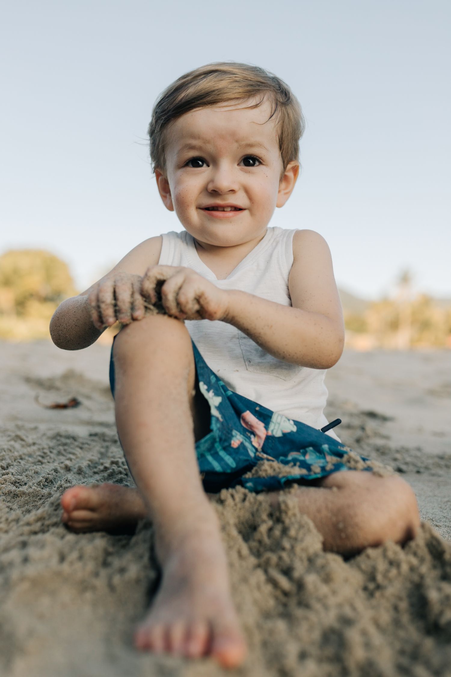 Smiling toddler enjoys tactile play in beach sand on a bright afternoon.