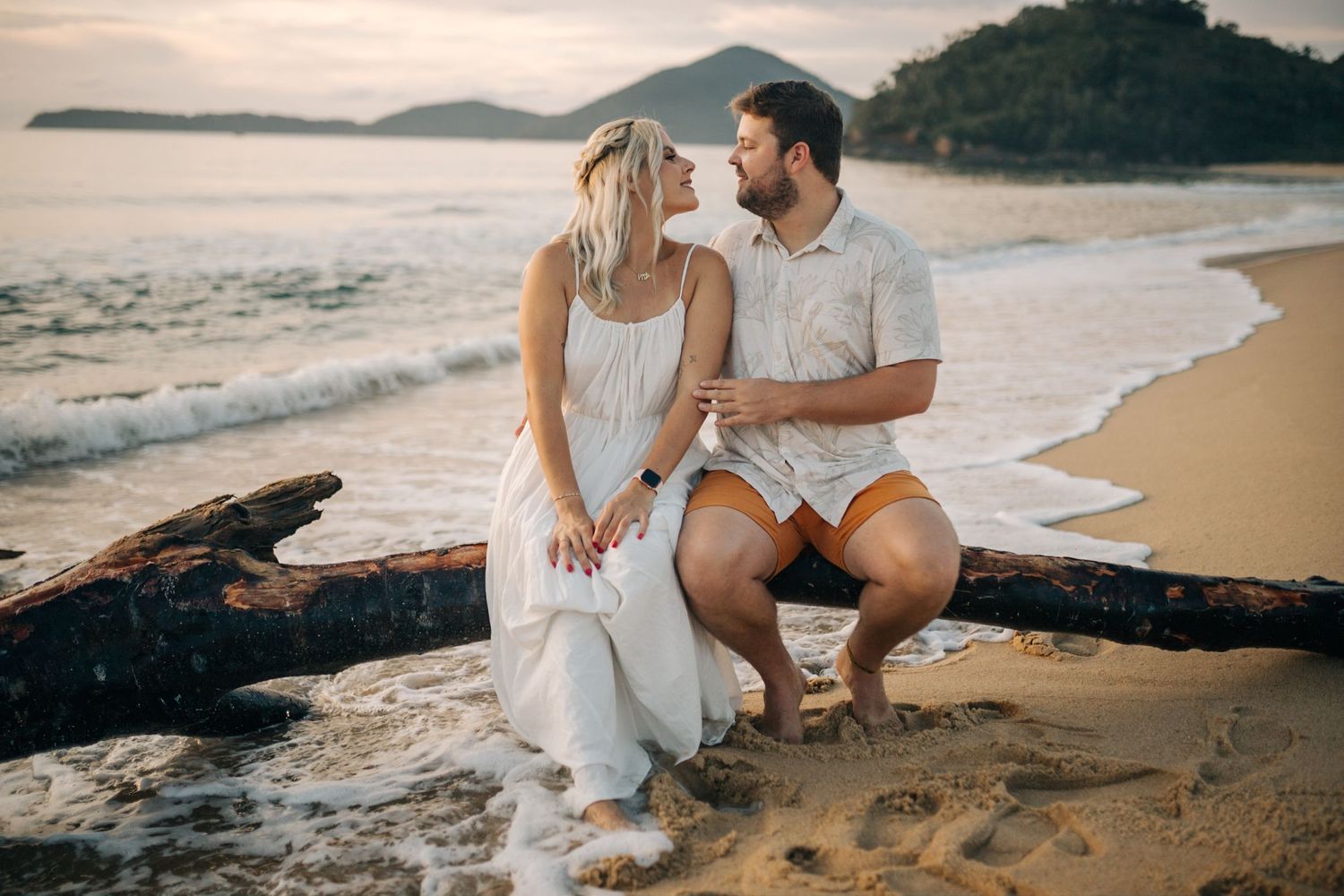 Two people share an intimate moment while sitting on a log at the beach during golden hour.