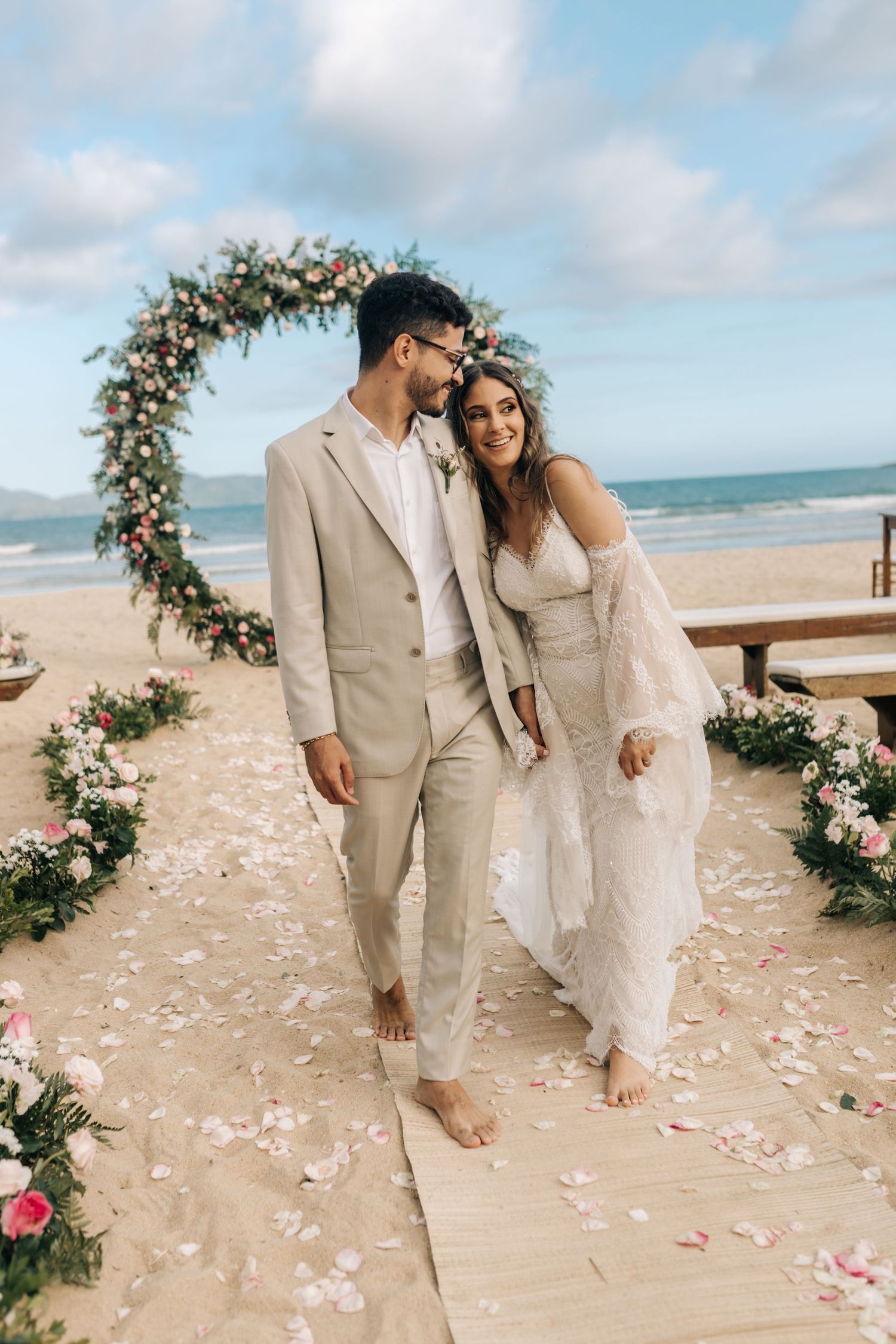 Wedding couple poses beneath a flower-adorned arch on a coastal boardwalk.