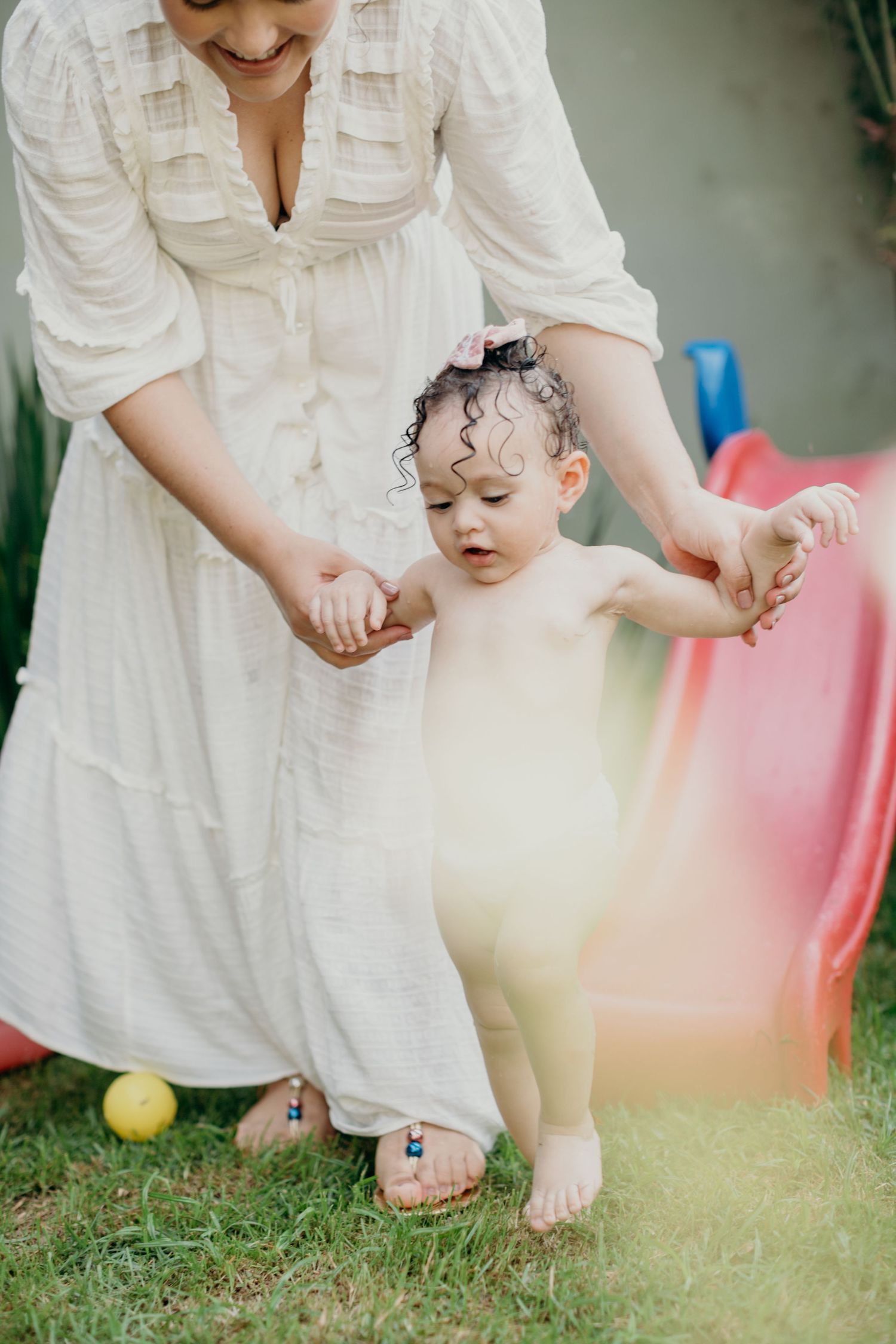 Person in white dress helping small child play with outdoor toys on grass.