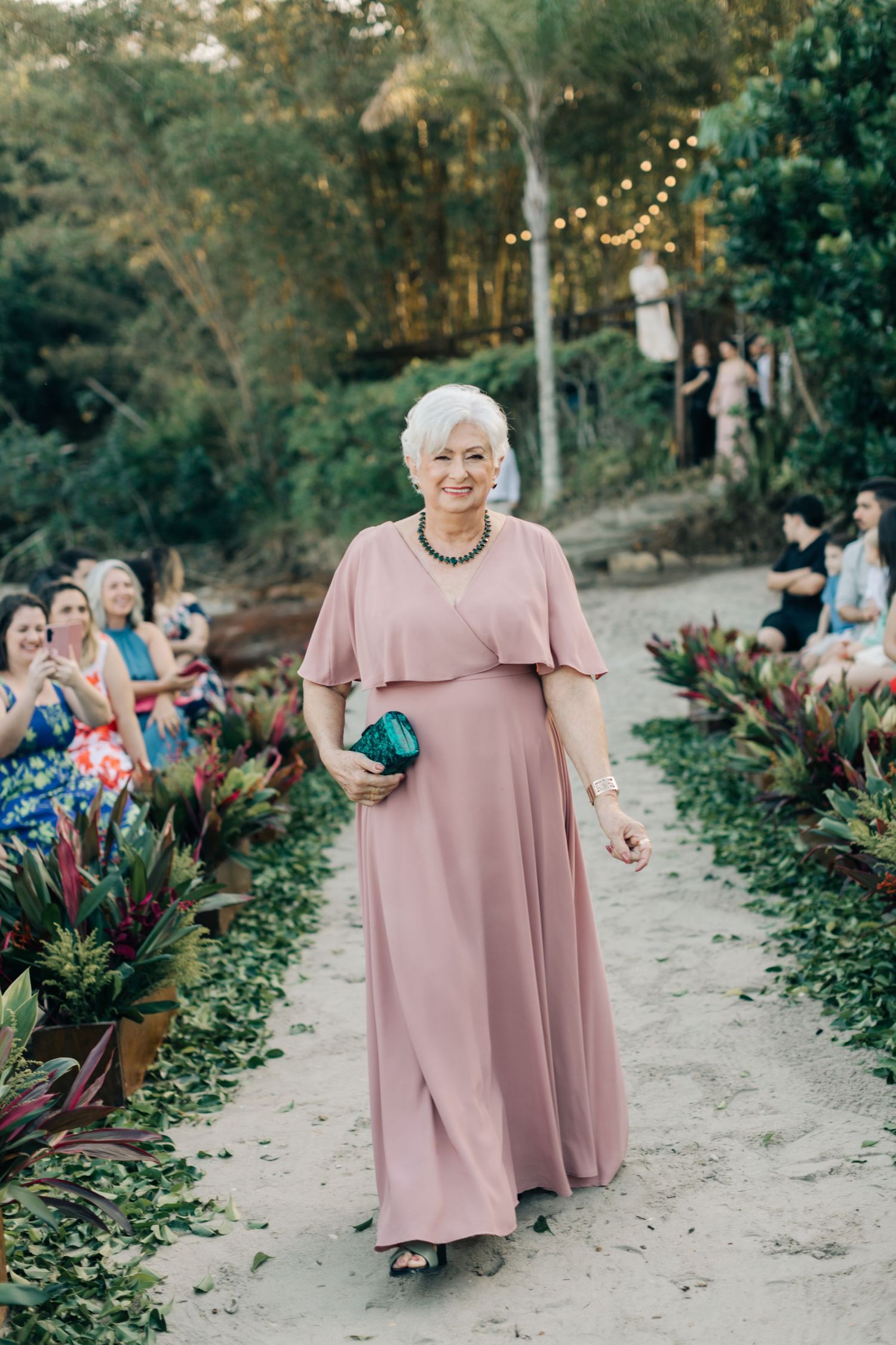 Wedding attendant in dusty pink maxi dress walks down garden path lined with tropical plants and string lights.