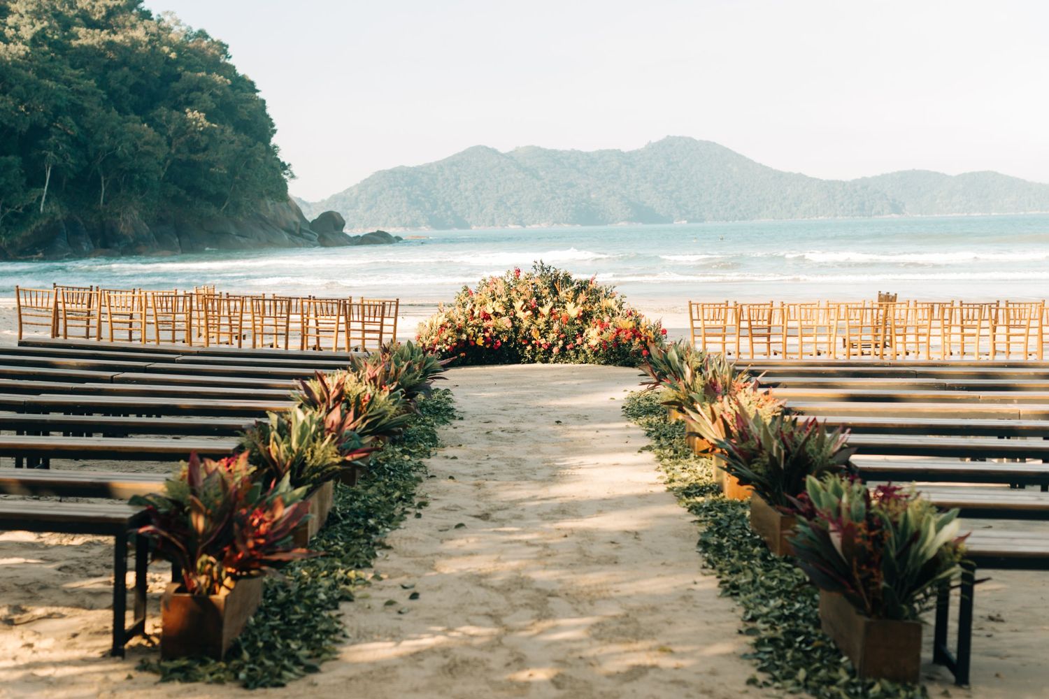 Beach wedding ceremony setup with wooden chairs and floral aisle decorations facing the ocean.