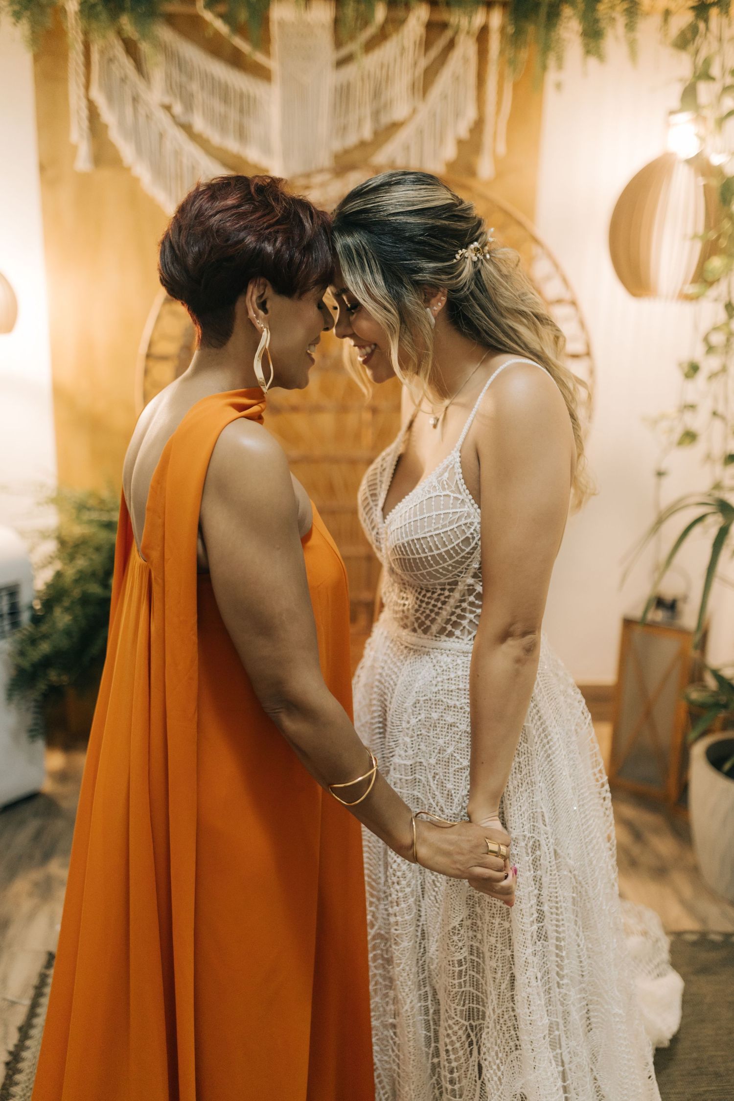 Intimate wedding moment with couple in orange dress and white lace gown sharing tender embrace against decorative backdrop.